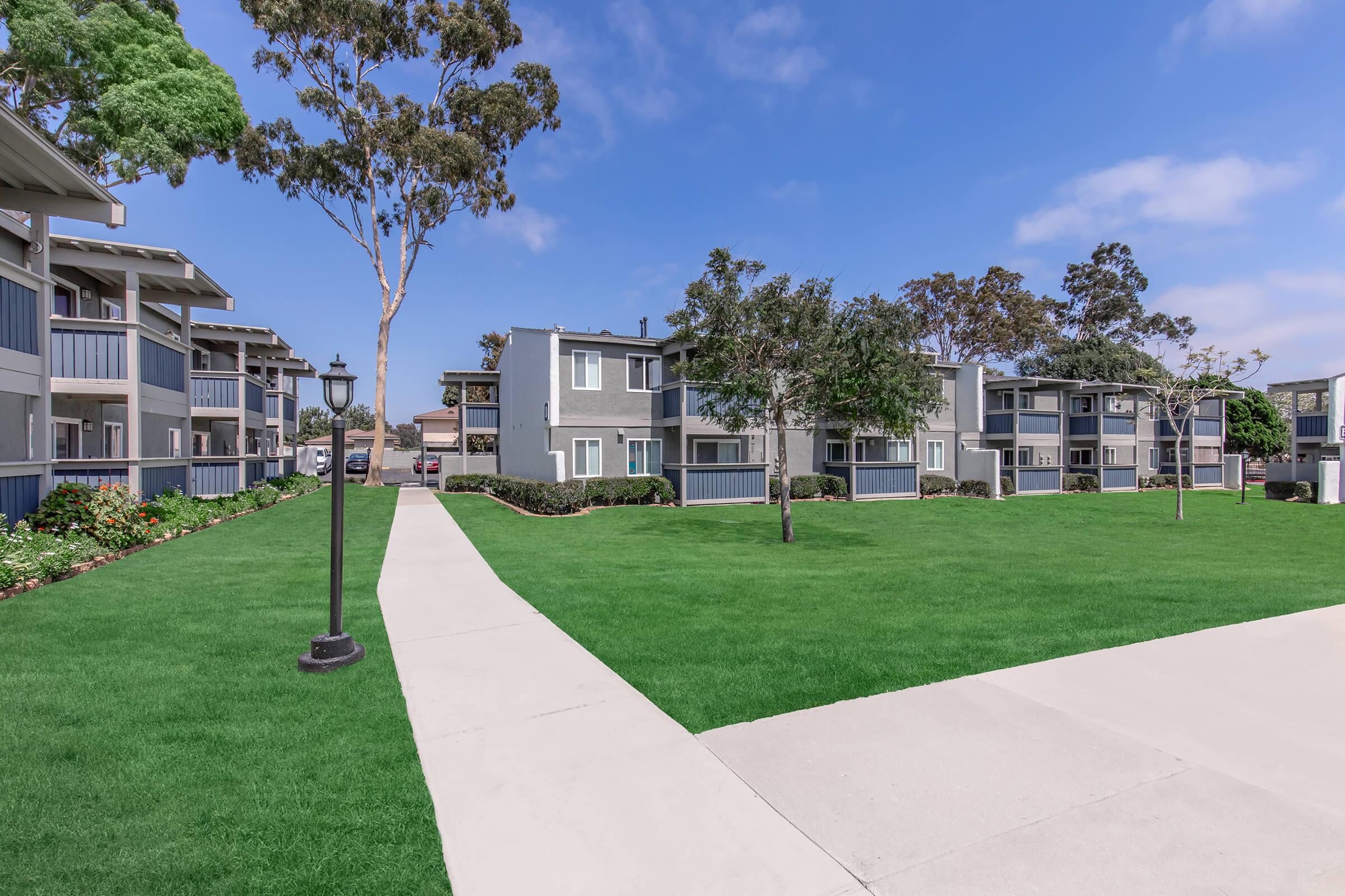 A peaceful residential area featuring two-story apartment buildings set amidst well-maintained lawns and trees. A paved walkway leads through the green space, with light posts adding to the inviting atmosphere under a clear blue sky.