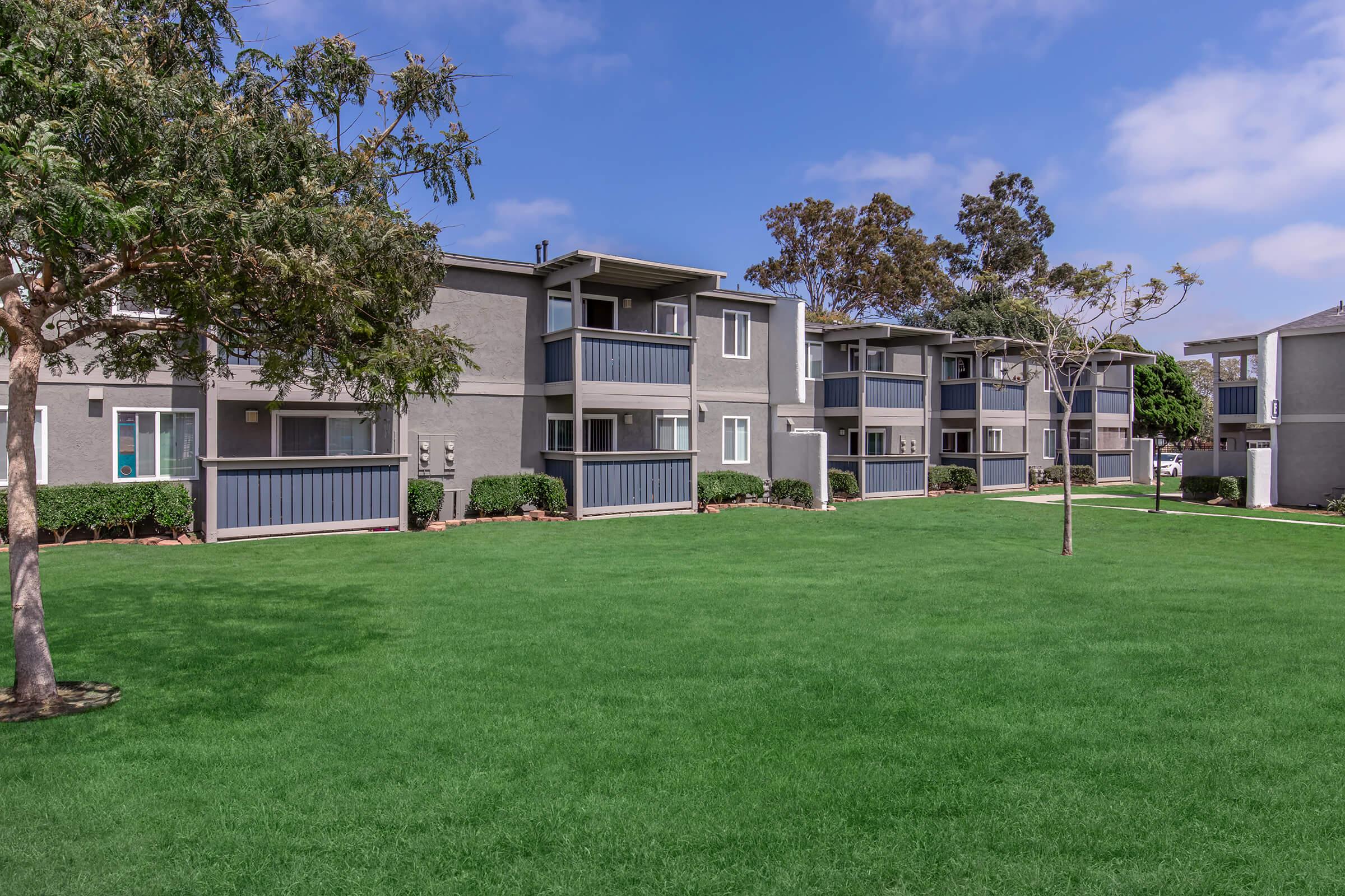 A view of a residential apartment complex featuring two-story buildings with balconies, set against a bright blue sky. Lush green grass and a few trees are visible in the foreground, creating a welcoming outdoor space.