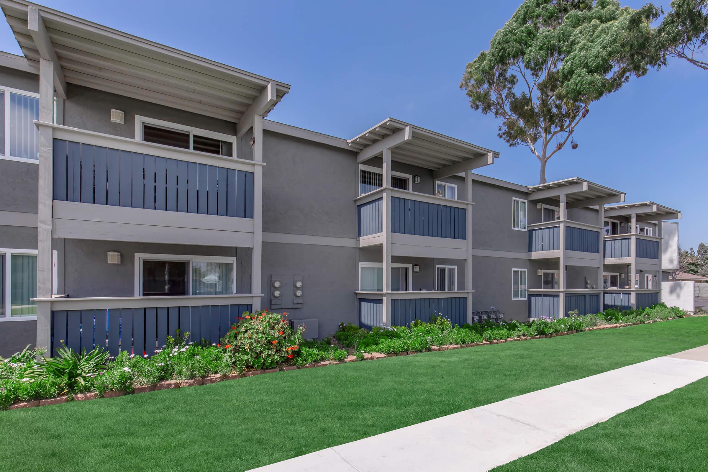 A modern apartment complex featuring multiple units with balconies, surrounded by lush green landscaping and a clear blue sky. The building has a gray exterior with blue accents and well-maintained flower beds along a paved walkway.