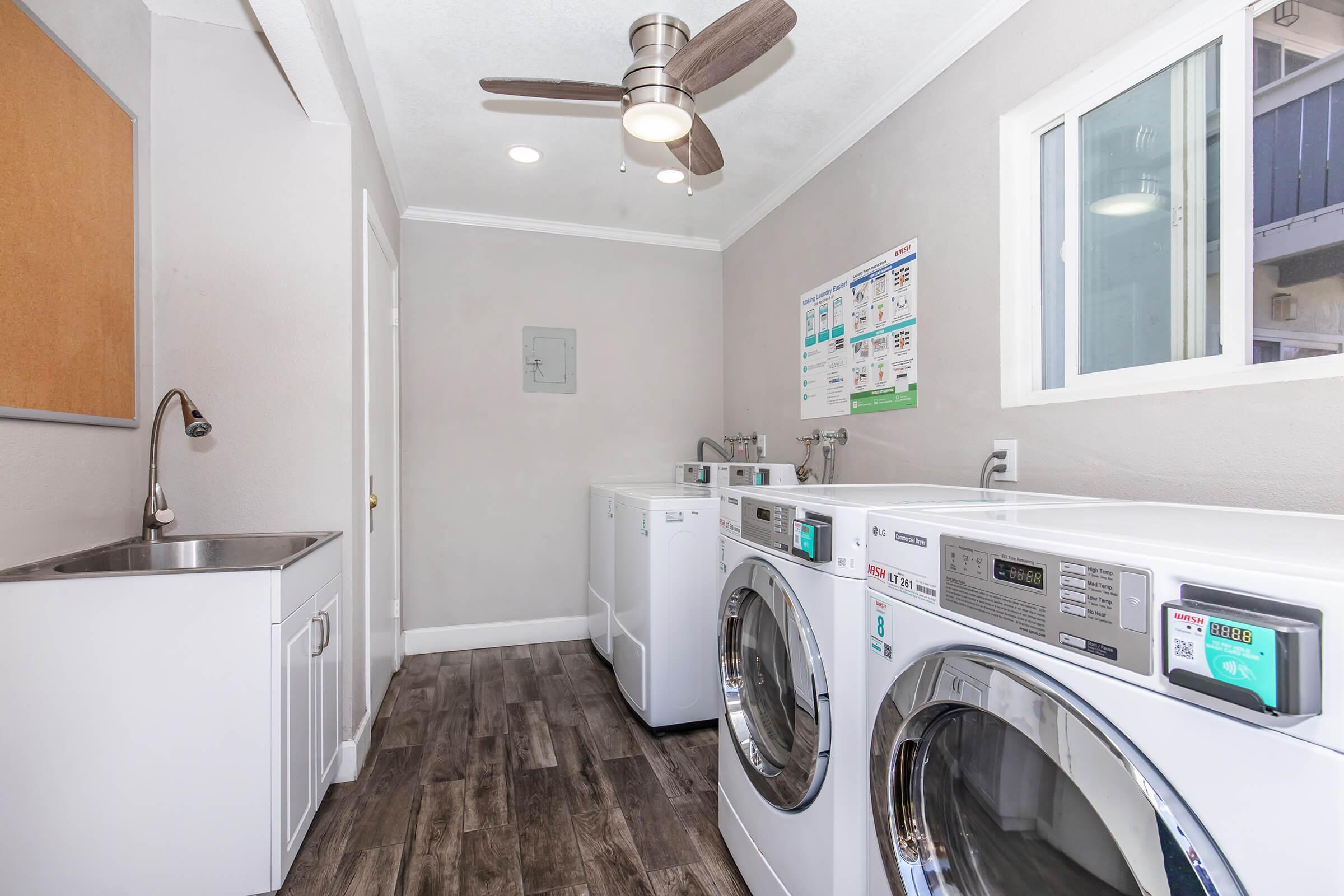 A clean and modern laundry room featuring multiple washing machines and dryers. There's a utility sink, a ceiling fan, and a window providing natural light. The walls are painted light gray, and there's a bulletin board with information on the wall. The floor is made of dark wood-like tiles.