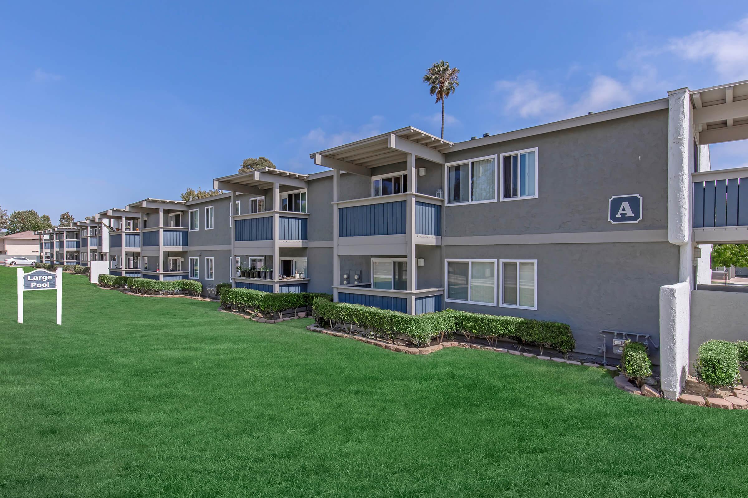 A row of modern apartment buildings with balconies, set on a well-maintained grassy area. The structures have gray exteriors and blue accents. A palm tree is visible beside the buildings. A sign labeled "Lawn Post" stands in the foreground, indicating the layout of the property. Bright blue sky completes the scene.