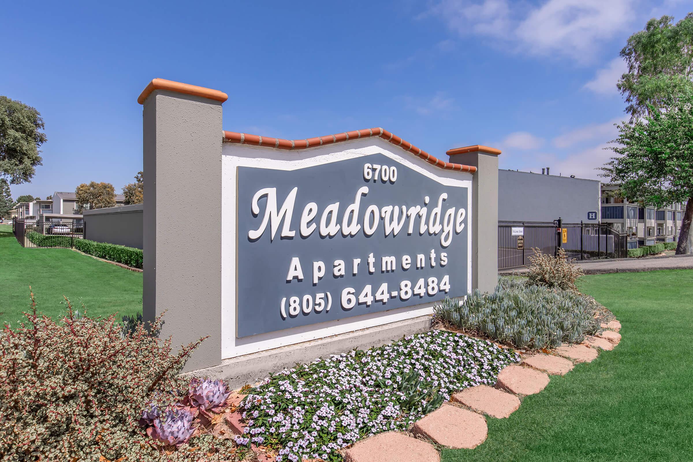 Sign for Meadowridge Apartments featuring the address 6700, phone number (805) 644-8484, surrounded by landscaped grass and flower beds. The background includes blue skies and trees, adding to the welcoming atmosphere of the apartment complex.