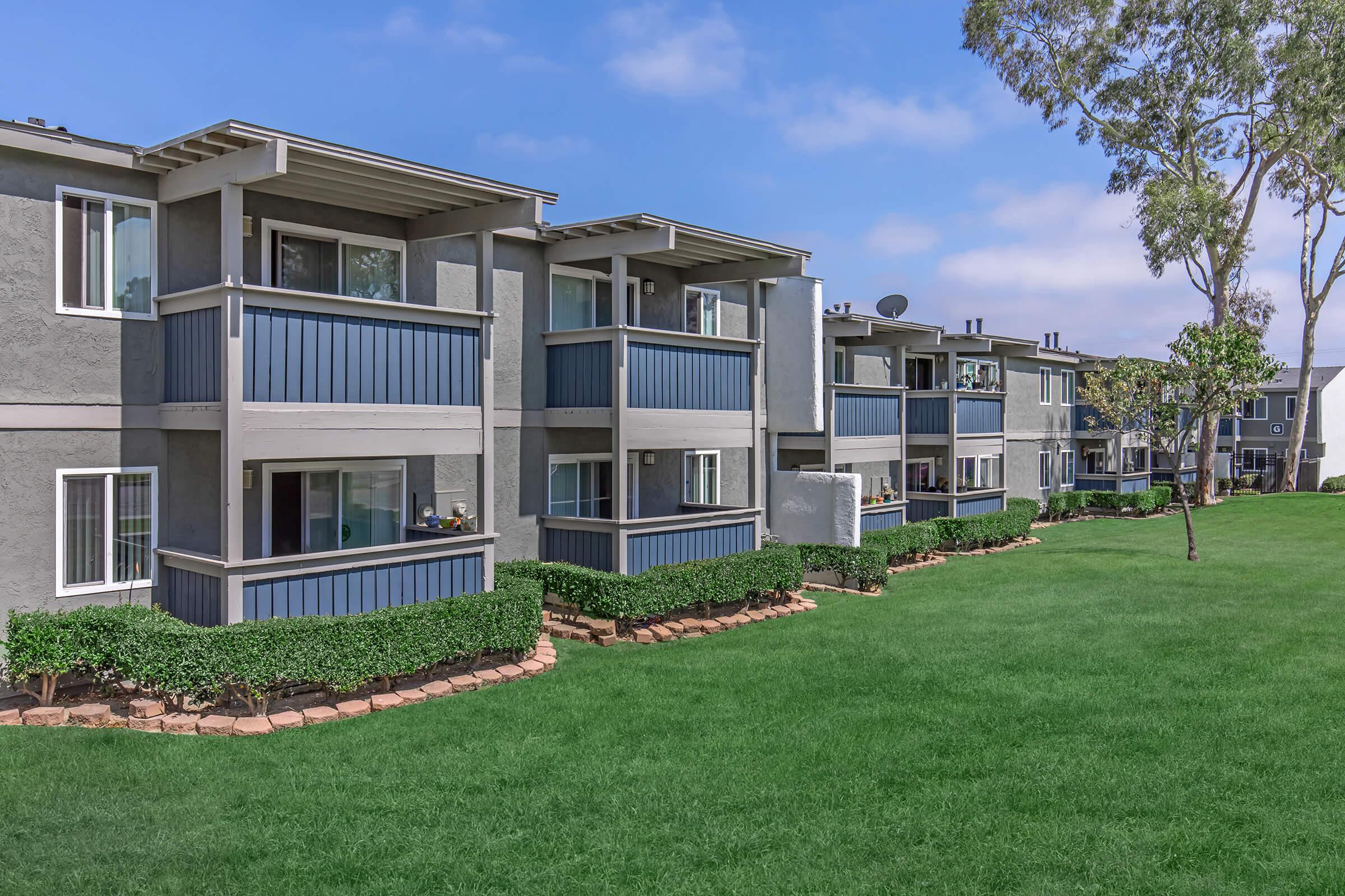 Row of modern apartment buildings with blue and gray exteriors, featuring balconies and well-maintained green lawns in front. The sky is partly cloudy, adding a serene atmosphere to the residential area.