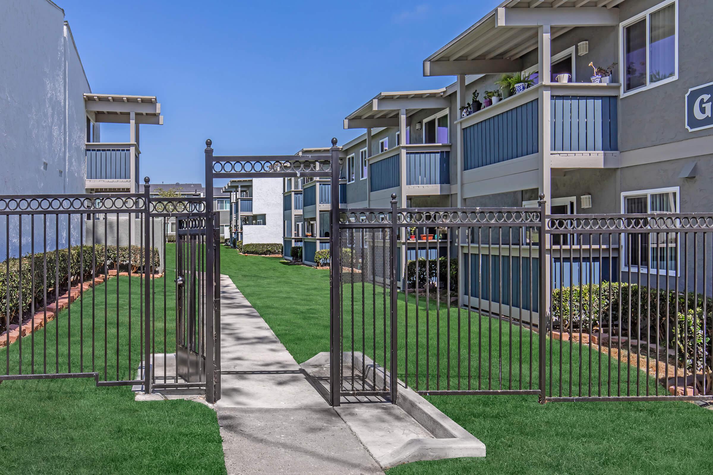 A landscaped area with a gated entrance leading to a pathway. On either side, there are two multi-story apartment buildings featuring balconies and blue accents. Lush green grass surrounds the pathway, creating a welcoming outdoor space. Clear blue skies are visible above.