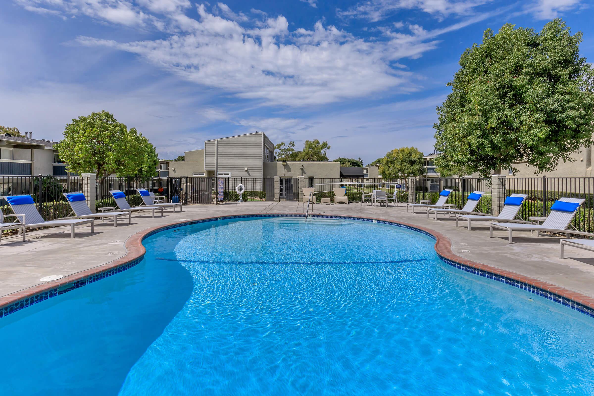 A clear, inviting swimming pool surrounded by lounge chairs with blue cushions. Lush green trees are nearby, and modern apartment buildings can be seen in the background. The sky is bright blue with scattered clouds, creating a relaxing and sunny atmosphere perfect for leisure.