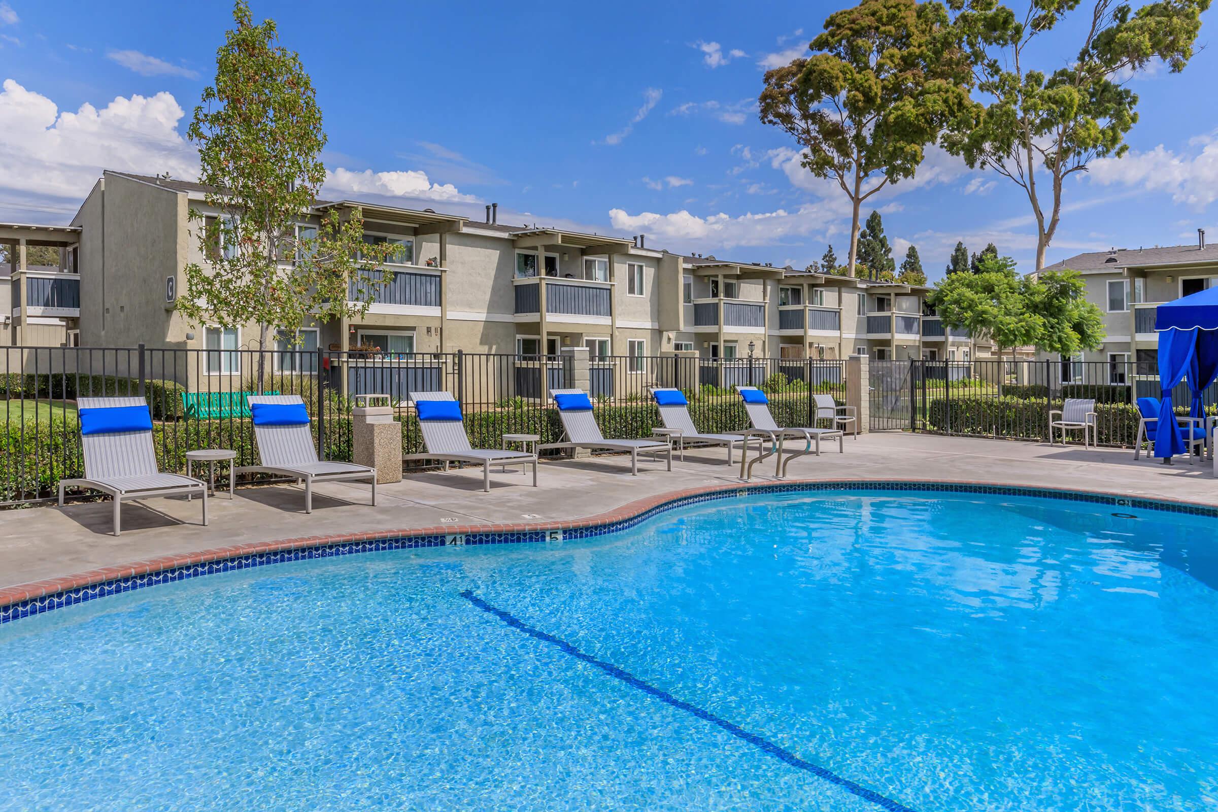 A sparkling blue swimming pool surrounded by lounge chairs under partly cloudy skies. In the background, there are apartment buildings with green landscaping and trees. The scene captures a relaxing outdoor space perfect for leisure and enjoyment.