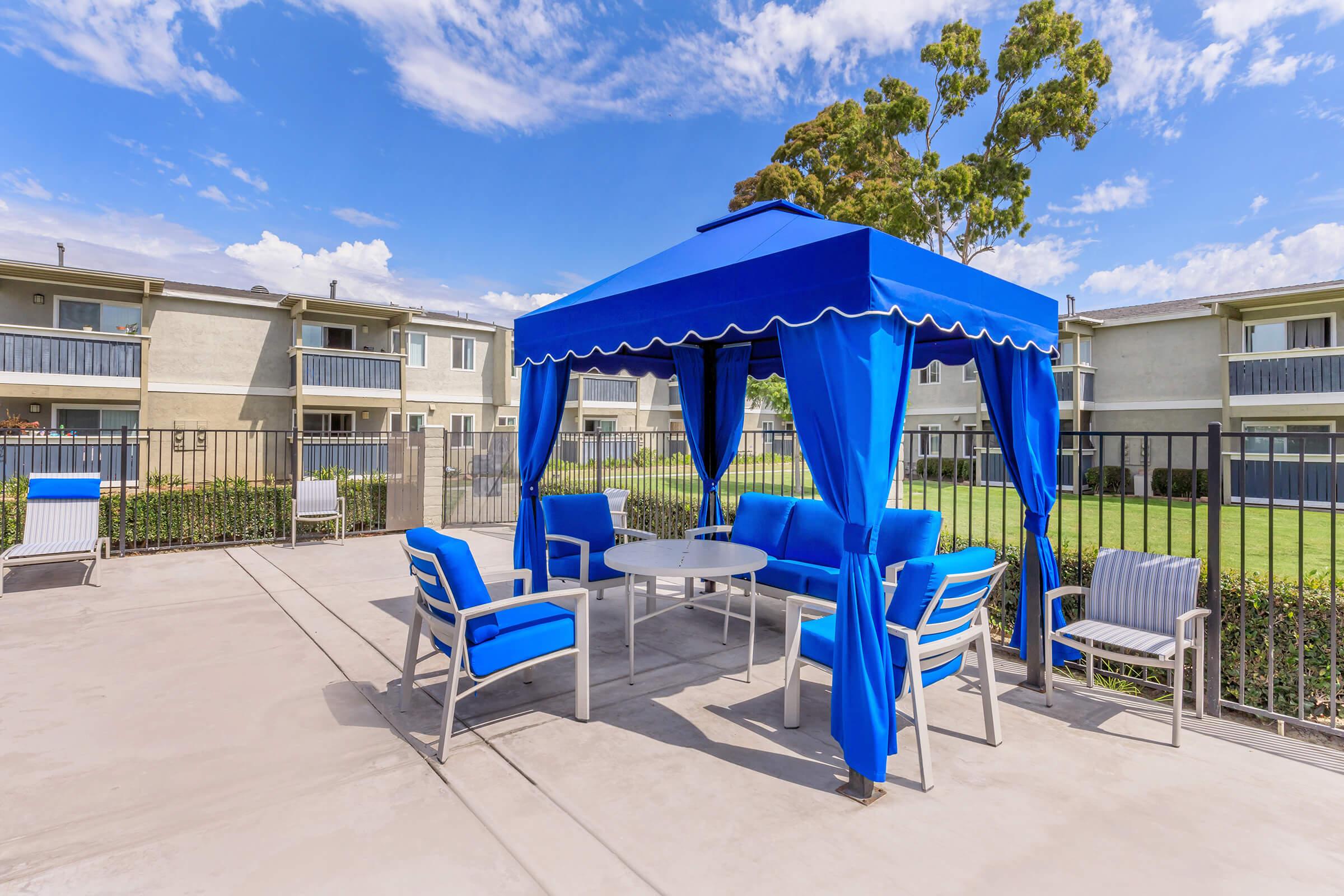 A vibrant blue canopy with seating arranged around a round table, set in a residential outdoor area. The space features well-maintained grass and buildings in the background, with a blue sky dotted with clouds, creating a welcoming and relaxing atmosphere.