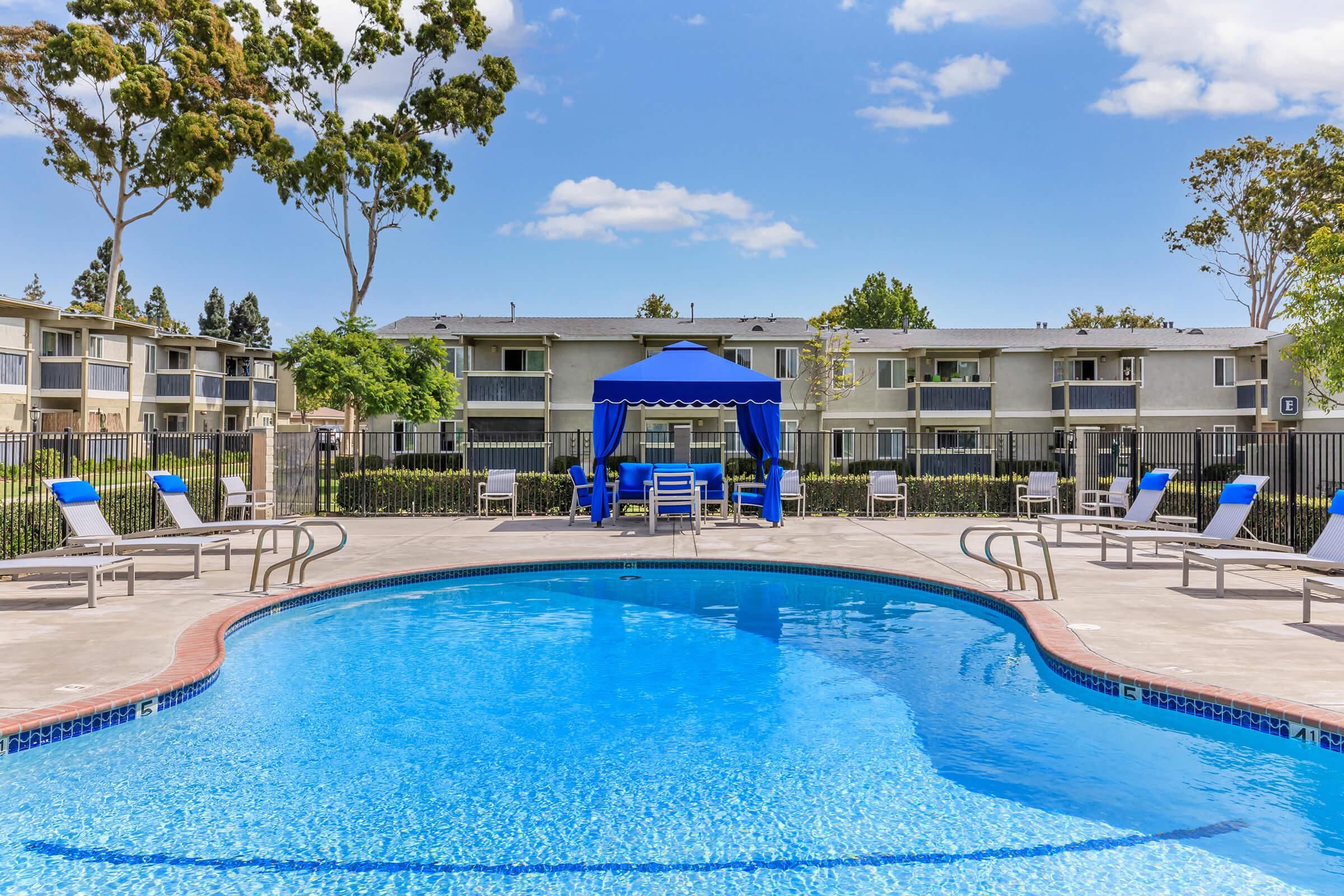 A serene swimming pool surrounded by lounge chairs and a blue canopy. The pool is beautifully clear, with a backdrop of trees and residential buildings. The sky is bright blue with a few clouds, creating a relaxing outdoor atmosphere.