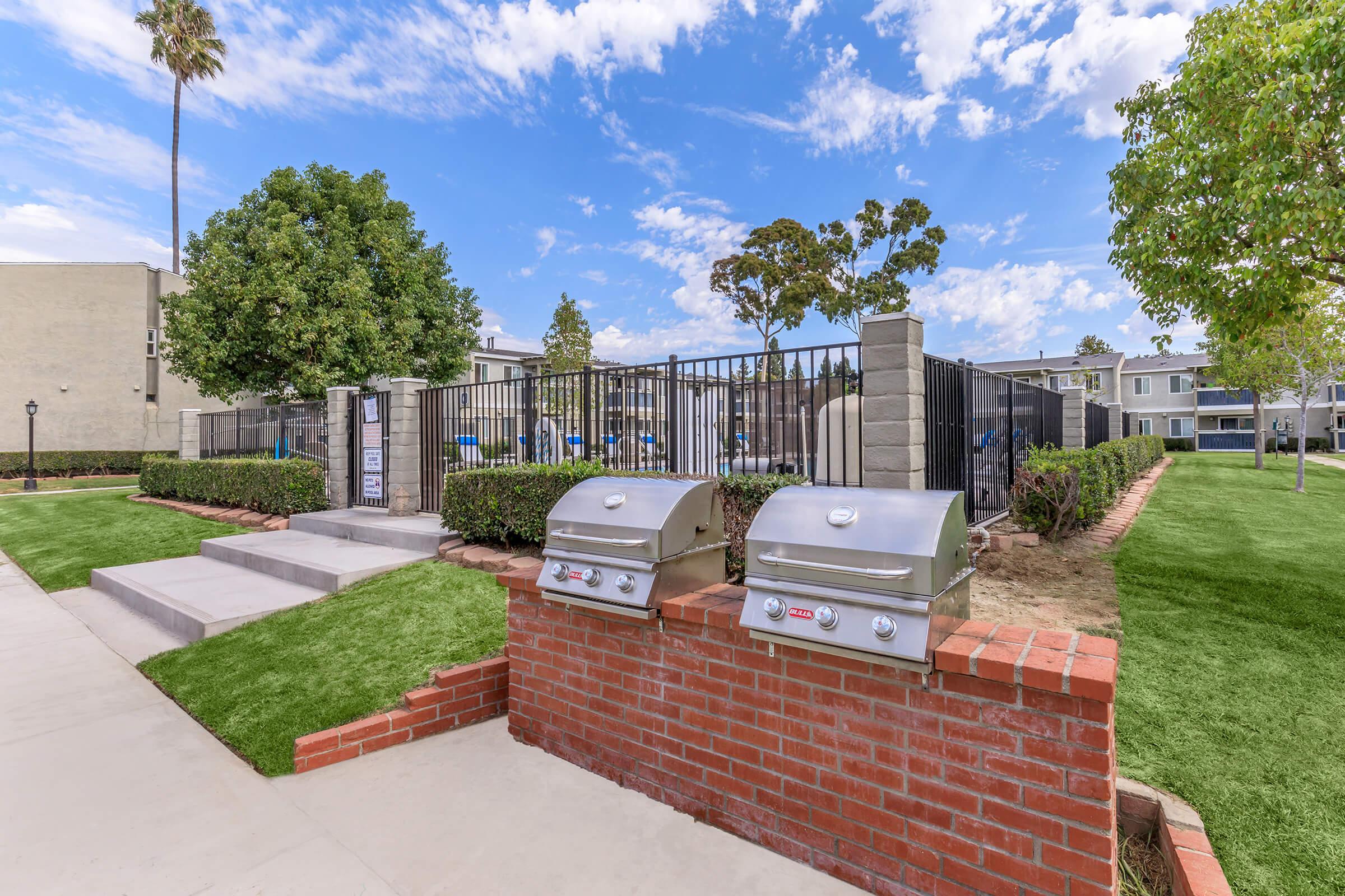 Outdoor barbecue area with two stainless steel grills situated on a brick platform. Surrounding greenery includes well-maintained grass and shrubs, with a clear blue sky and scattered clouds above. In the background, a fenced pool area and several apartment buildings are visible.