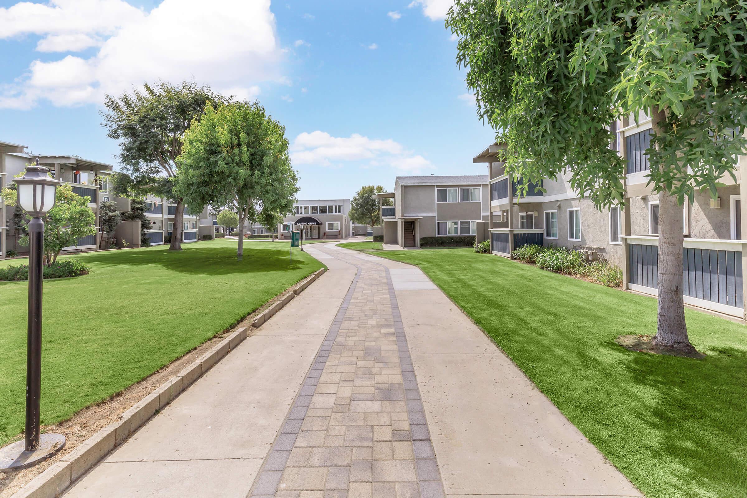 A well-maintained pathway winding through a green landscape, flanked by trees and modern apartment buildings. The sky is bright with fluffy clouds, creating a serene and inviting atmosphere. Well-kept lawns add to the overall appeal of the outdoor space.