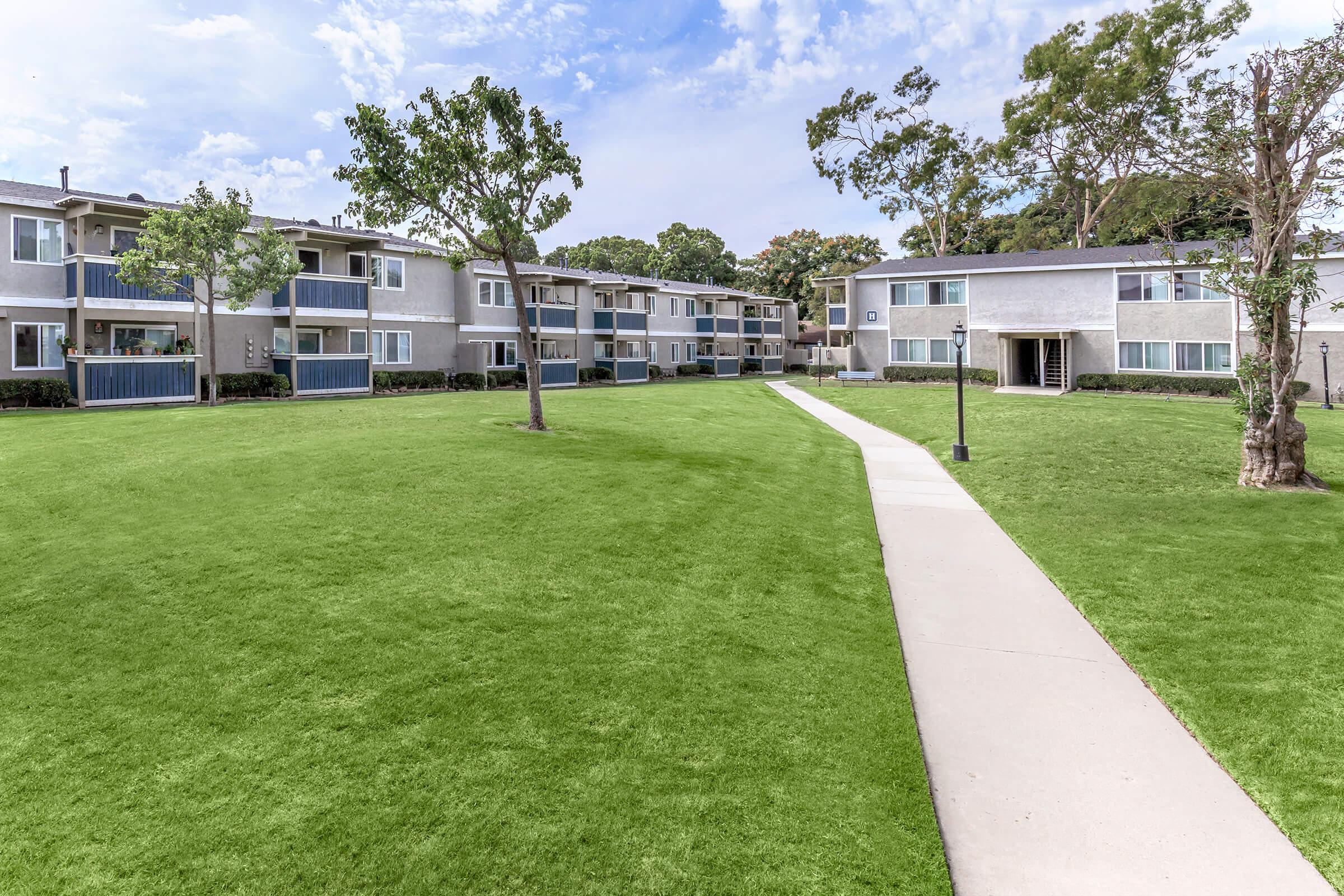 A well-maintained apartment complex with two-story buildings surrounding a manicured lawn. A smooth concrete pathway runs through the green grass, leading to entrances of the apartments. Trees are strategically placed throughout the area, and the sky is partly cloudy, adding to the pleasant atmosphere.