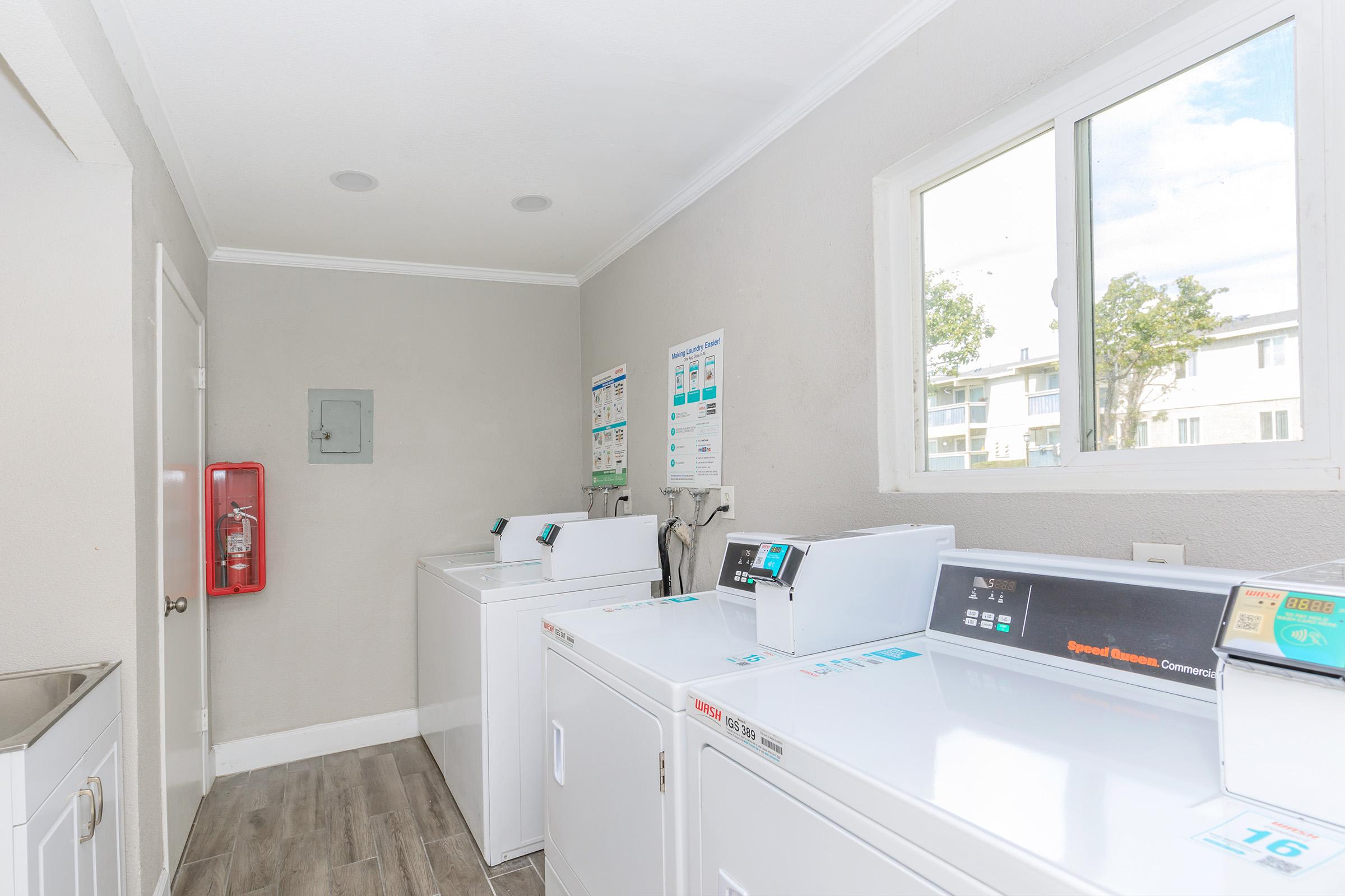 A laundry room featuring several white washing machines and dryers, with a window providing natural light. The walls are painted a light gray, and there is a fire extinguisher mounted on the wall. Various informational posters are displayed near the machines.