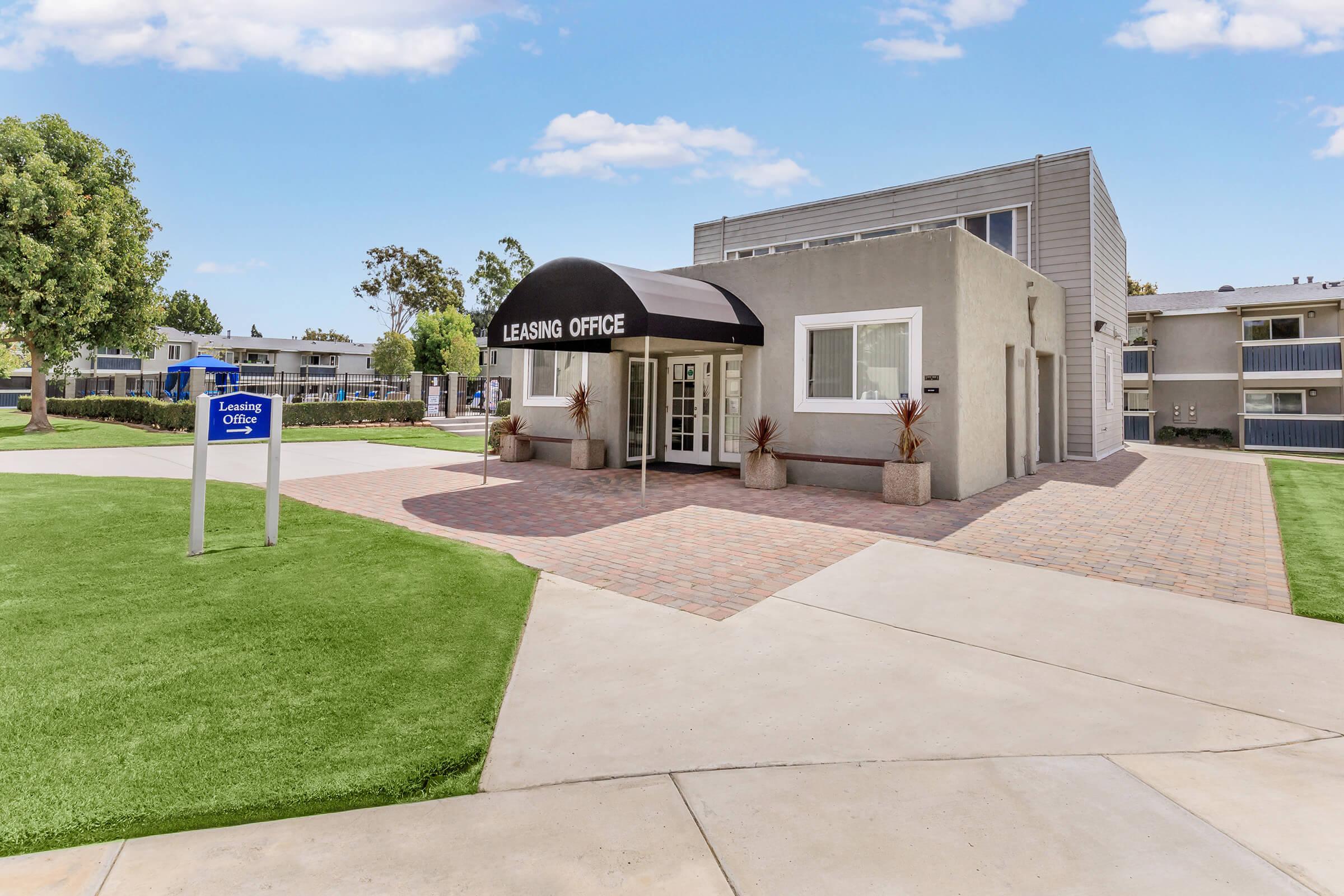 A modern leasing office with a black awning labeled "Leasing Office." The building features a gray exterior and is surrounded by well-maintained grassy areas and pathways. A sign stands nearby indicating the leasing office location.