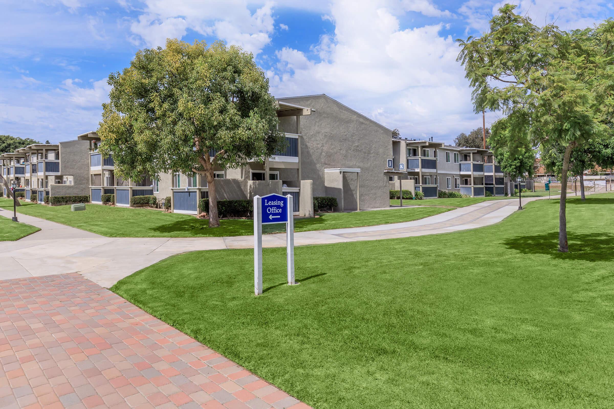 A well-maintained apartment complex with several two-story buildings surrounded by lush green lawns and trees. A sign for the leasing office is prominently displayed in front, along with paved walkways that lead through the landscaped grounds under a partly cloudy blue sky.