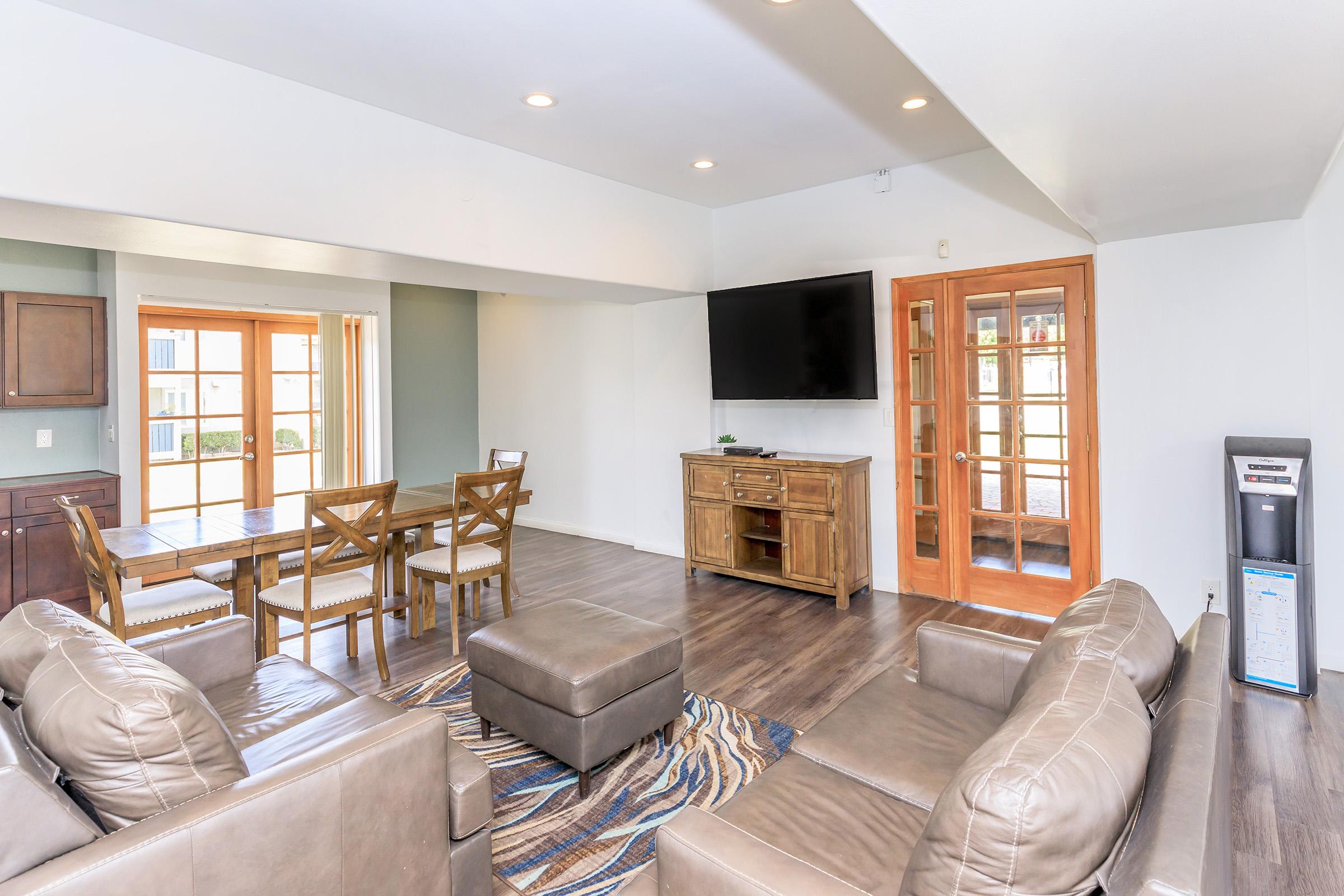 A spacious living area featuring a comfortable seating arrangement with brown leather sofas, a coffee table, and a wooden dining table with chairs. There’s a large flat-screen TV mounted on the wall and glass doors leading to an outdoor patio. Natural light fills the room, enhancing the modern decor.