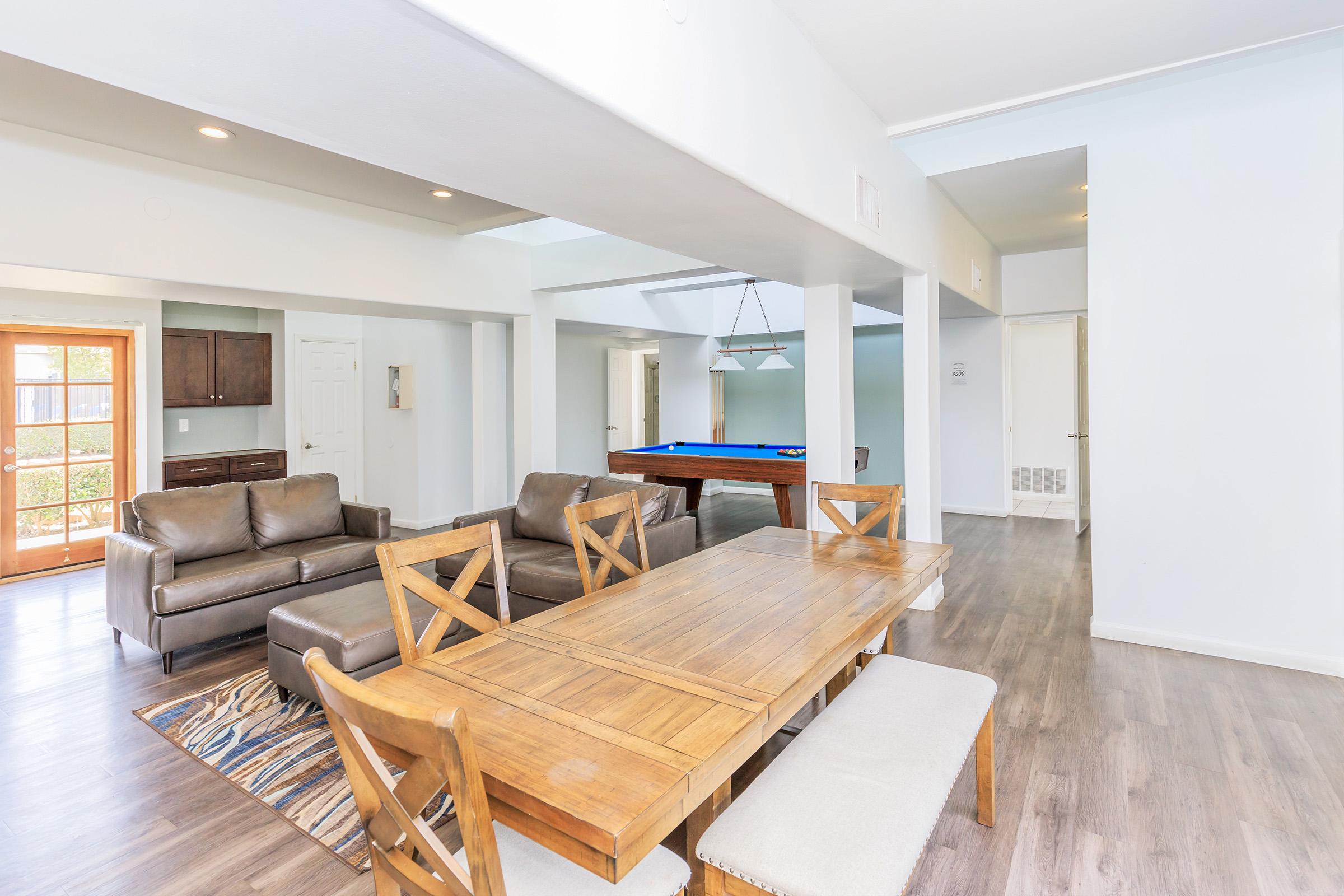 Interior view of a modern living space featuring a wooden dining table with chairs, two brown sofas, and a pool table in the background. The room has white walls, large windows allowing natural light, and a light wood floor. A doorway leads to the exterior area, enhancing the open feel of the space.