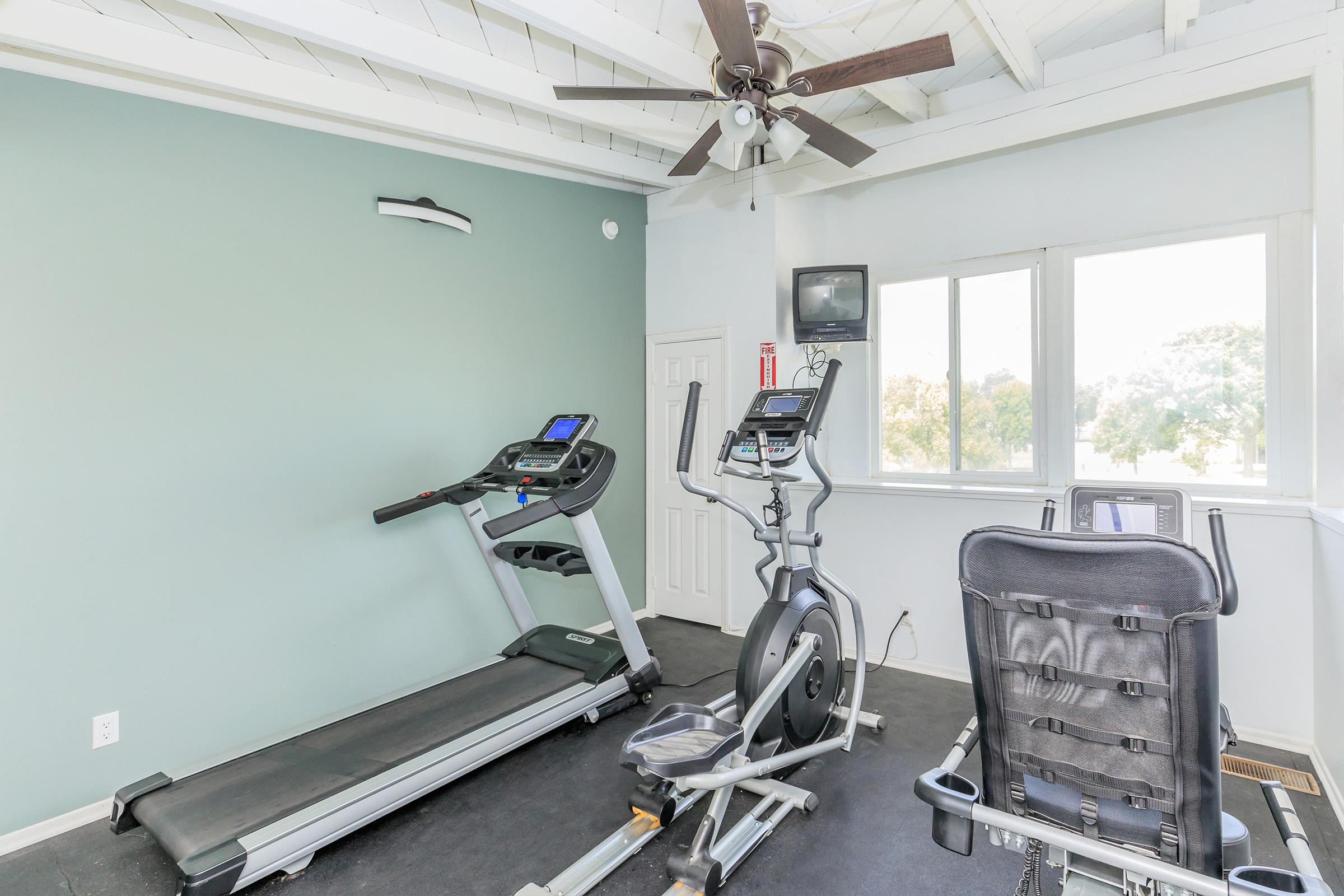 A fitness room featuring a treadmill, an elliptical machine, and a stationary bike. The room has a light green wall, ceiling fan, and large windows allowing natural light. A small TV is mounted on the wall, providing entertainment options while exercising.