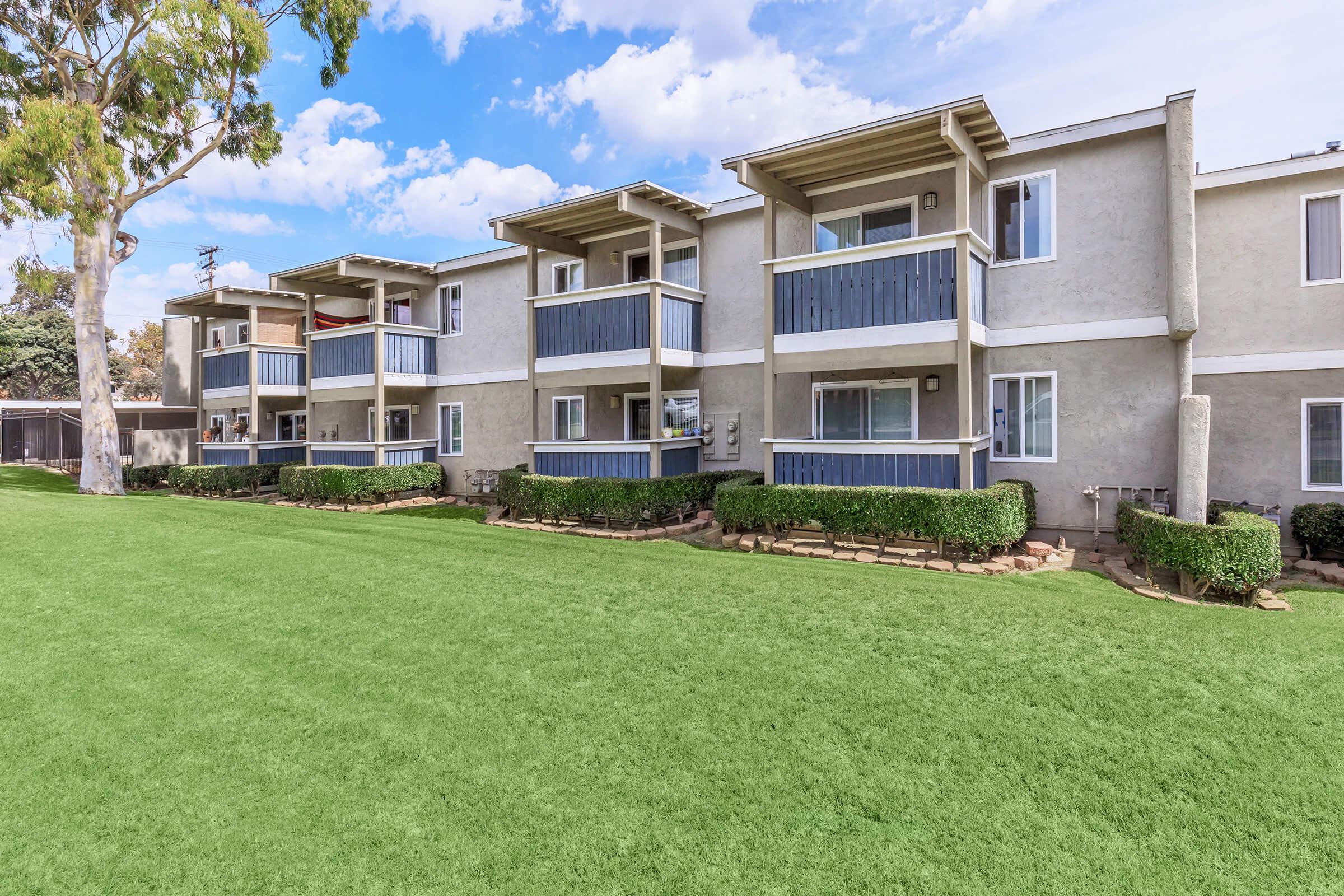 A row of modern apartment buildings with balconies, situated on a well-maintained green lawn. The sky is partly cloudy, and there are trees nearby, adding to the pleasant outdoor setting.