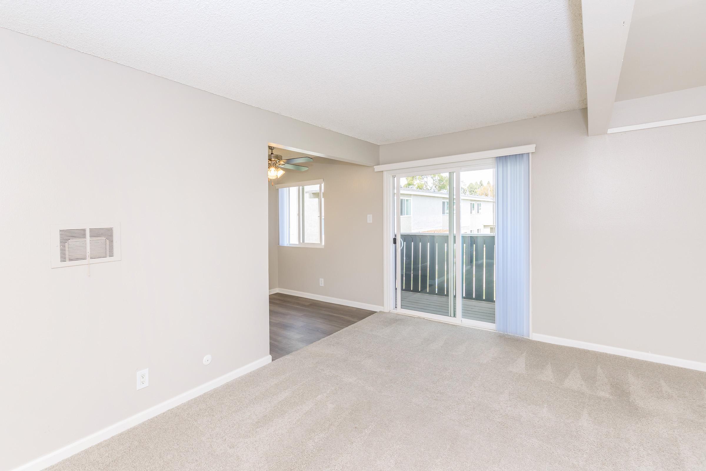Spacious interior of a light-colored living area with plush carpet, featuring a sliding glass door that opens to a balcony. A ceiling fan is visible, along with a window allowing natural light. The adjacent room is partially visible, enhancing the open layout of the space.