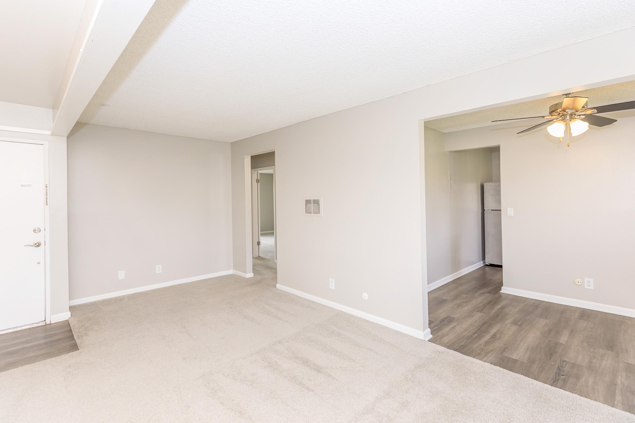 A spacious, well-lit living area featuring neutral-colored walls and carpet. To the left, a door leads to the entrance. There are two openings: one to a room and another to a kitchen area visible in the background. A ceiling fan hangs in the room, adding to the airy feel.