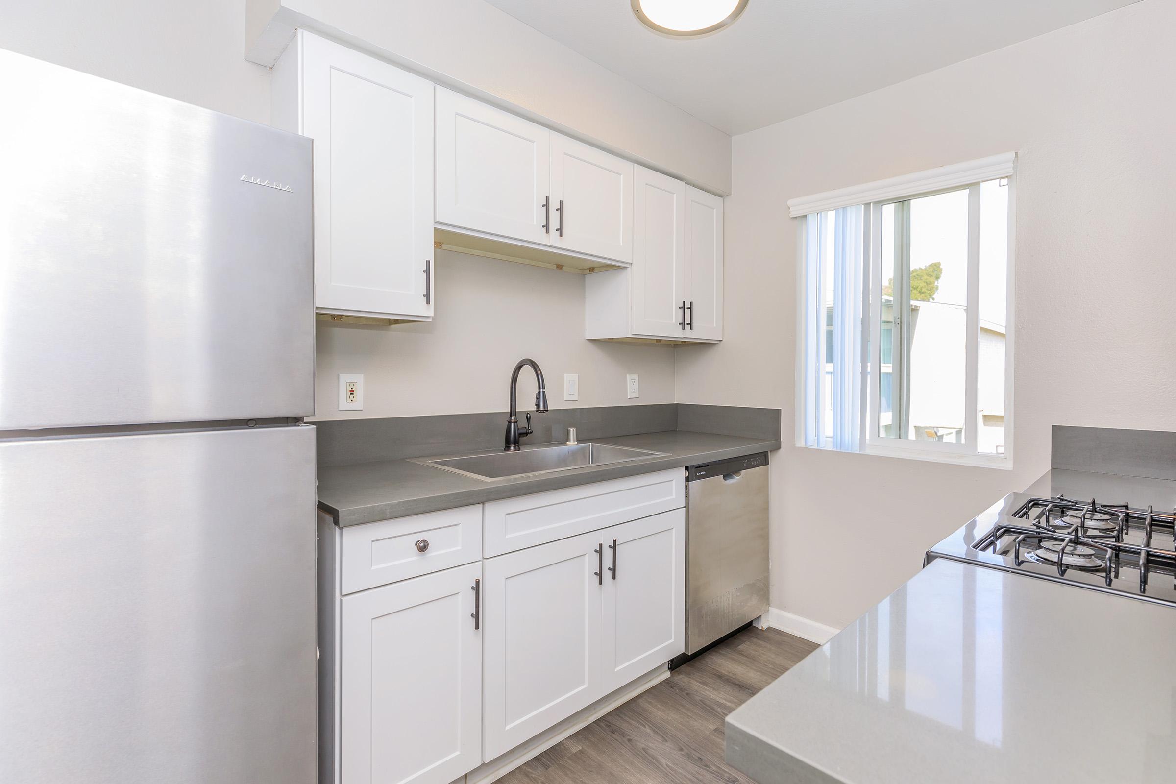 Modern kitchen featuring stainless steel appliances, including a refrigerator, sink, and a gas stove. The cabinets are white with a sleek design, and the countertop is gray. Natural light comes in through a window with vertical blinds. The flooring is a light wood finish, adding to the contemporary feel.