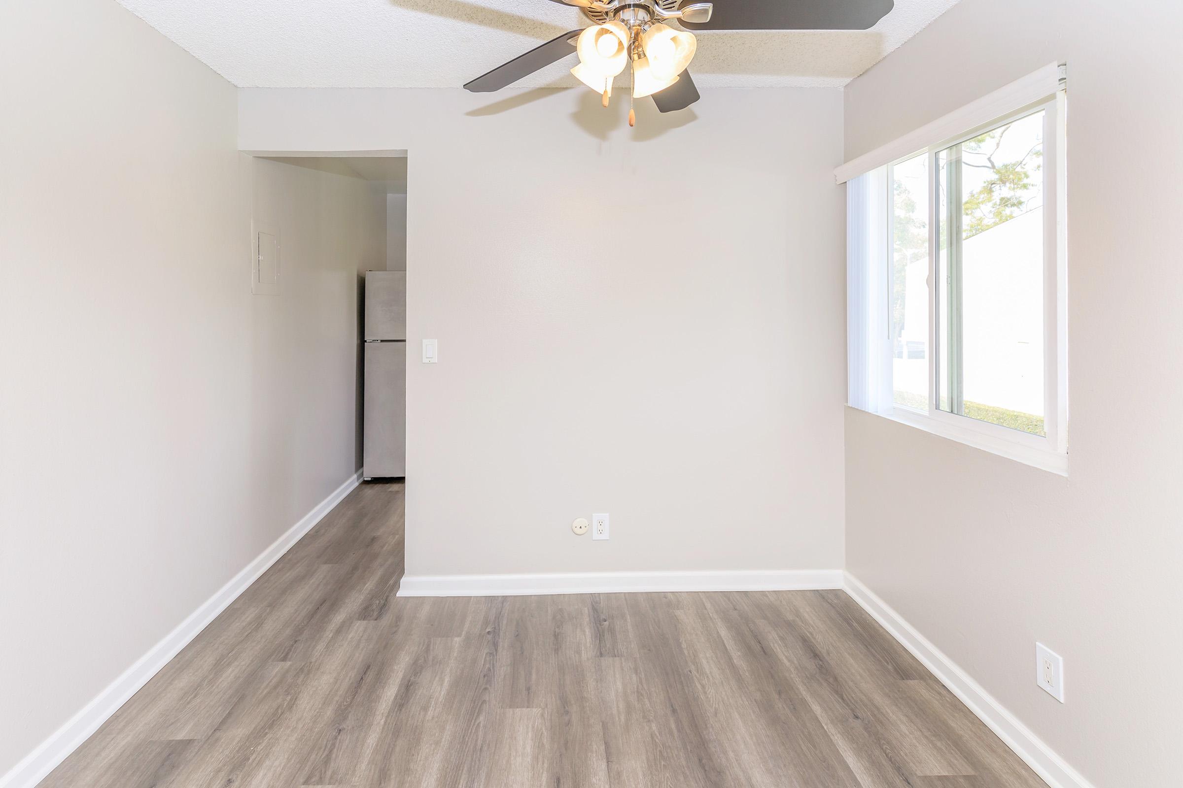 A spacious, empty room featuring a ceiling fan, light-colored walls, and modern laminate flooring. Natural light enters through a window on one side, while a doorway leads to a small kitchenette area visible in the background. The overall atmosphere is bright and inviting.