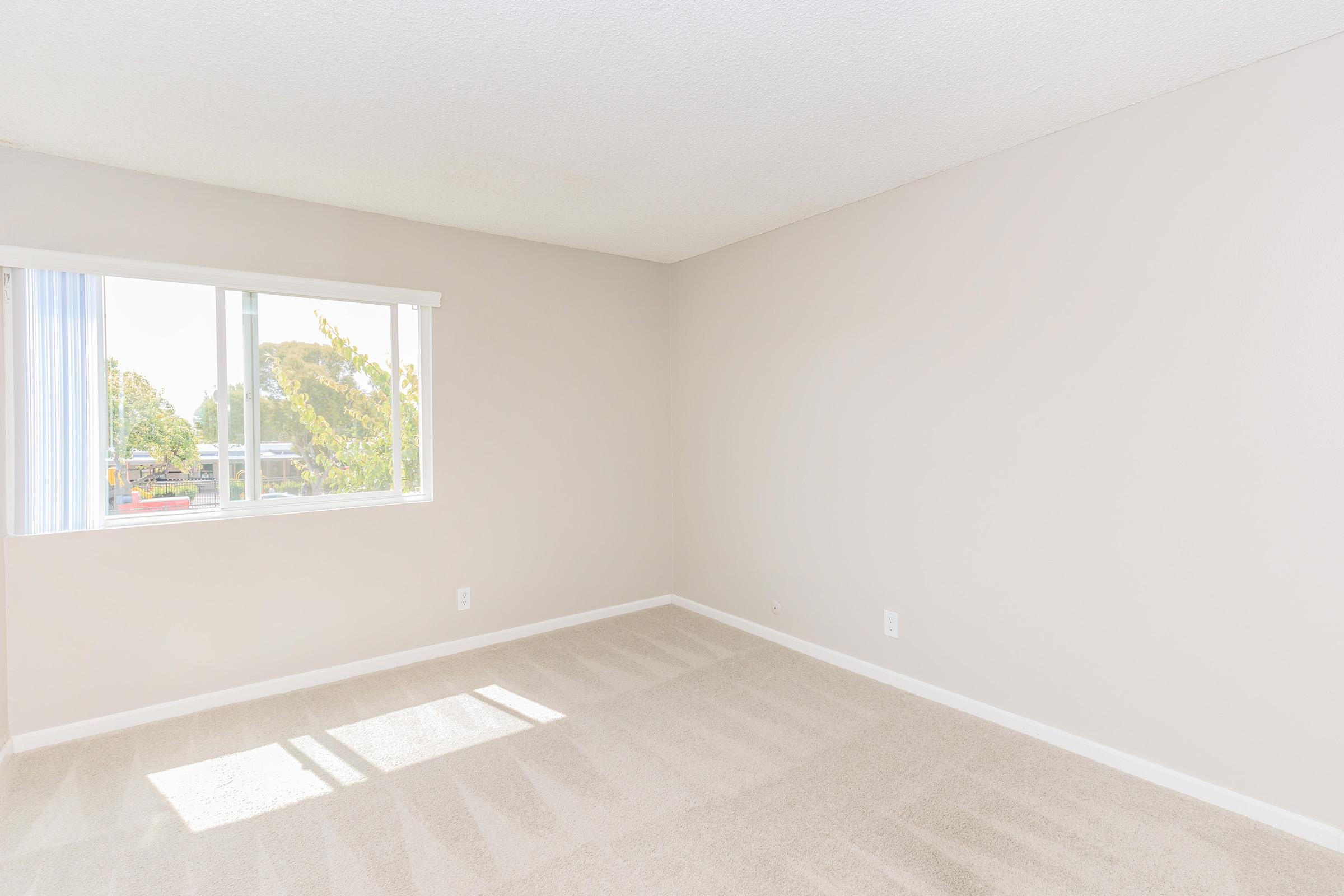 A bright, empty room with light beige walls and carpet. A large window lets in natural light, illuminating the space. Simple and clean design with no furniture, creating a minimalist atmosphere.