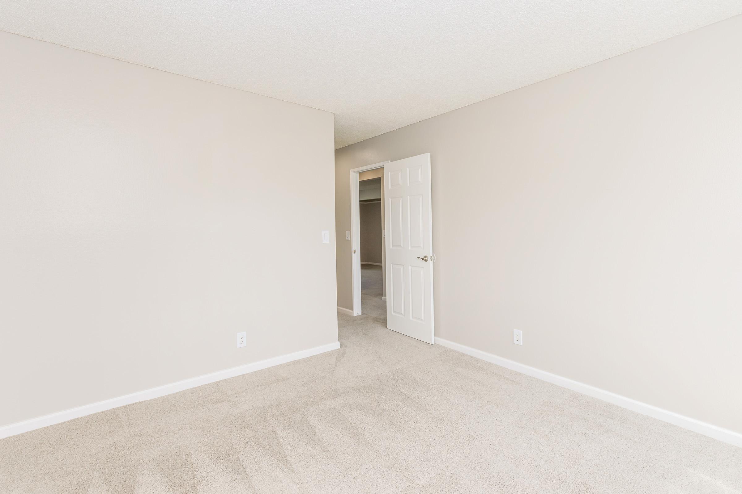 A well-lit, empty room with beige walls and light-colored carpet. A closed door is visible on the left, leading to another space, while an open doorway is seen in the background, suggesting access to additional rooms. The overall atmosphere is neutral and spacious.