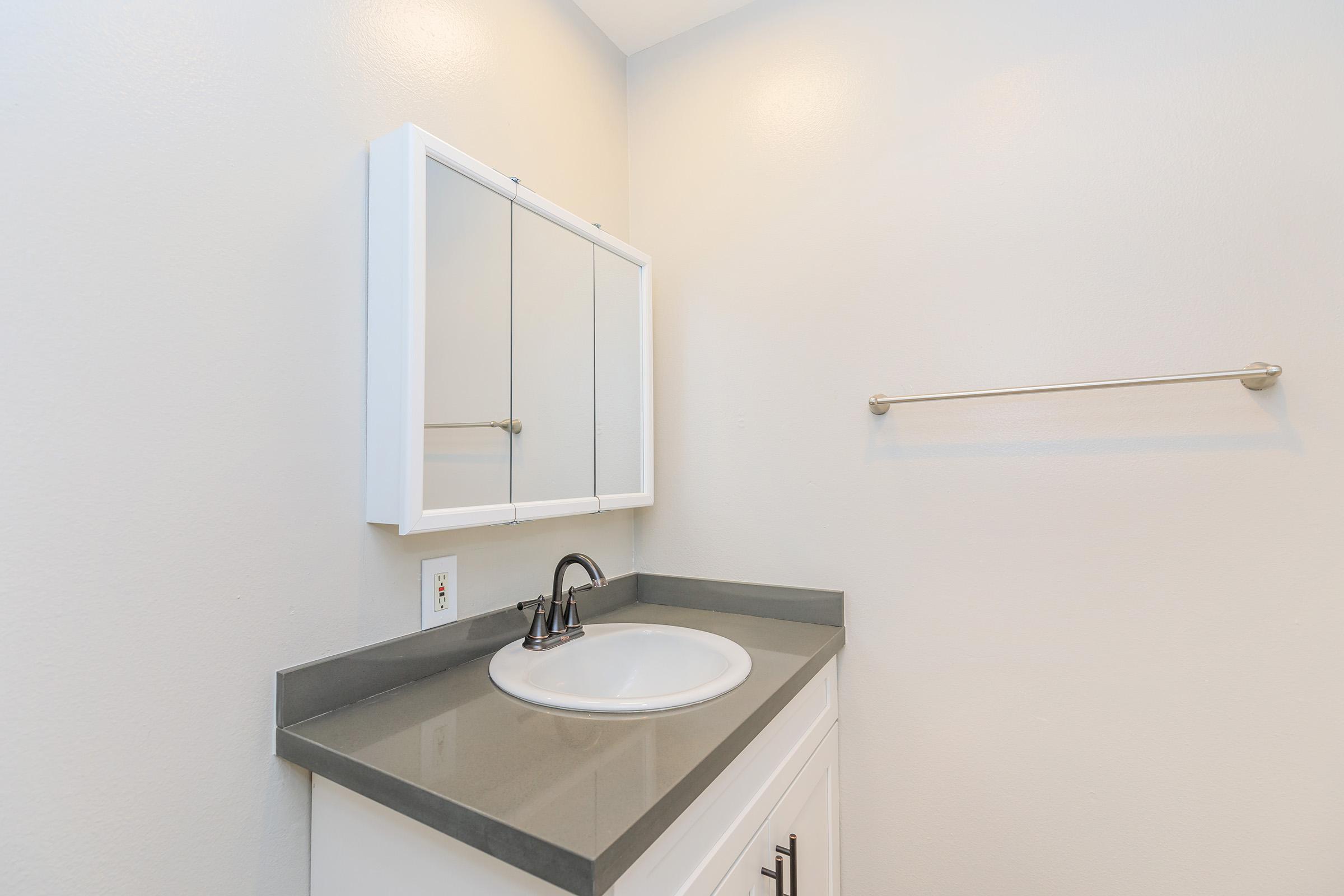 A modern bathroom sink area featuring a circular basin on a gray countertop, a white cabinet below, and a mirror with three panels above. The wall is a soft light color, and there is a towel bar mounted to the side.