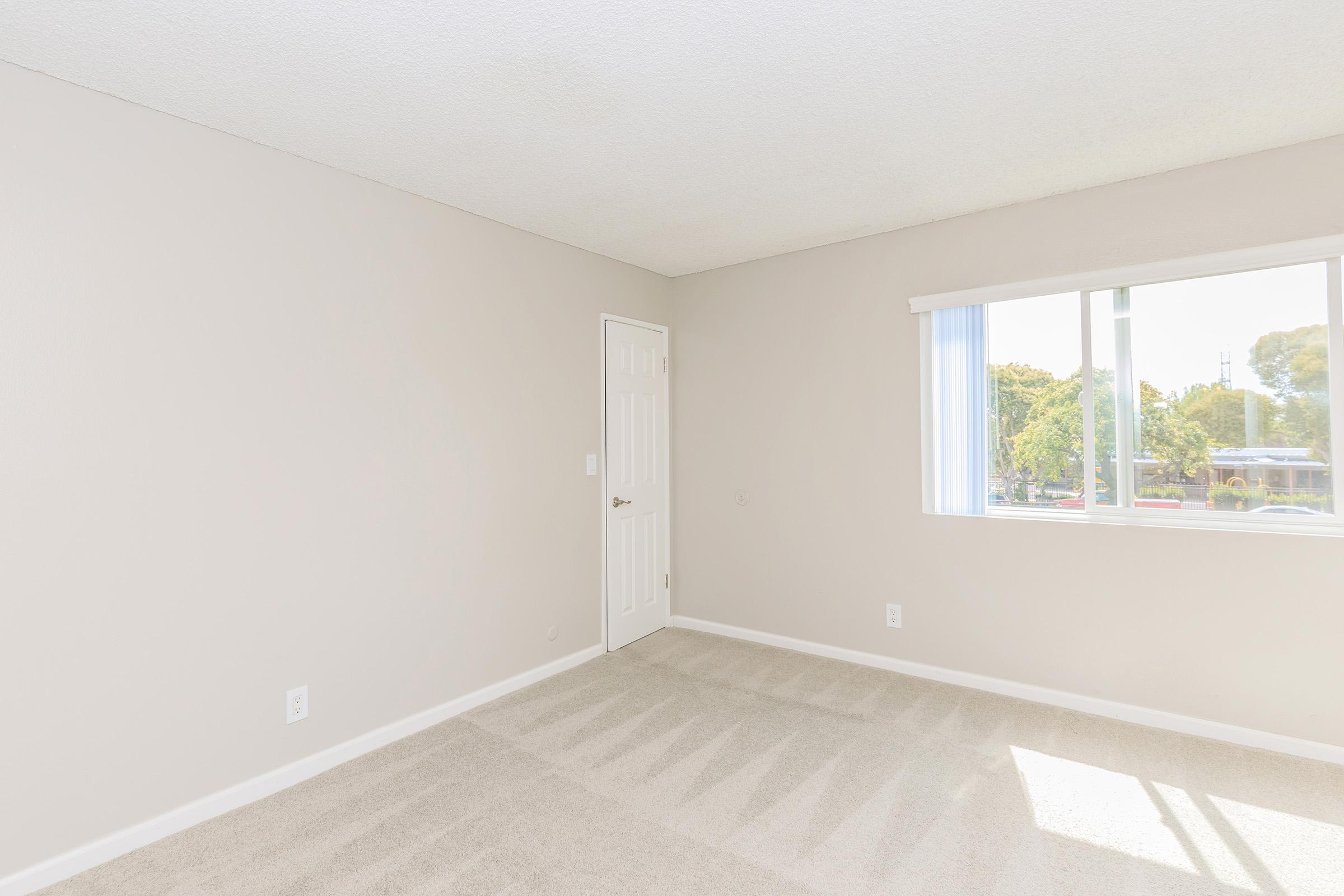 A well-lit, empty room with light beige walls and carpet. There is a white door on one side and a window with sheer curtains allowing natural light. The space appears clean and minimalistic, suitable for various uses such as a bedroom or office.