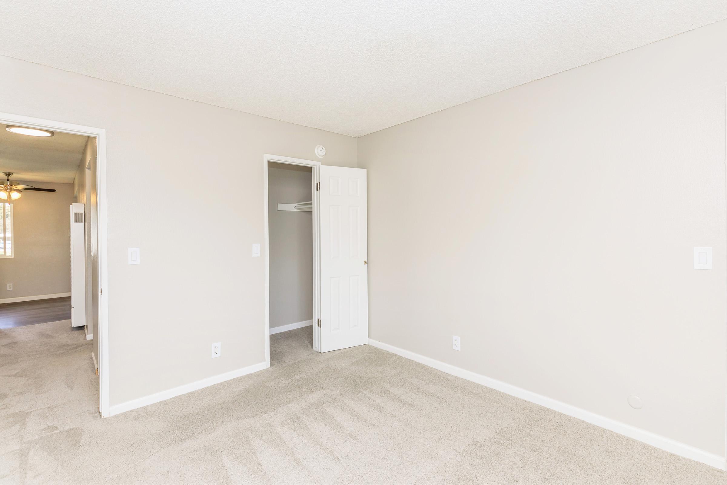 A well-lit, empty room with beige walls and light beige carpeting. It features a closed door leading to a closet on one side and an open doorway leading to another room on the opposite side. The ceiling has a textured finish, and there is a small wall outlet visible. Natural light enters from a window out of view.