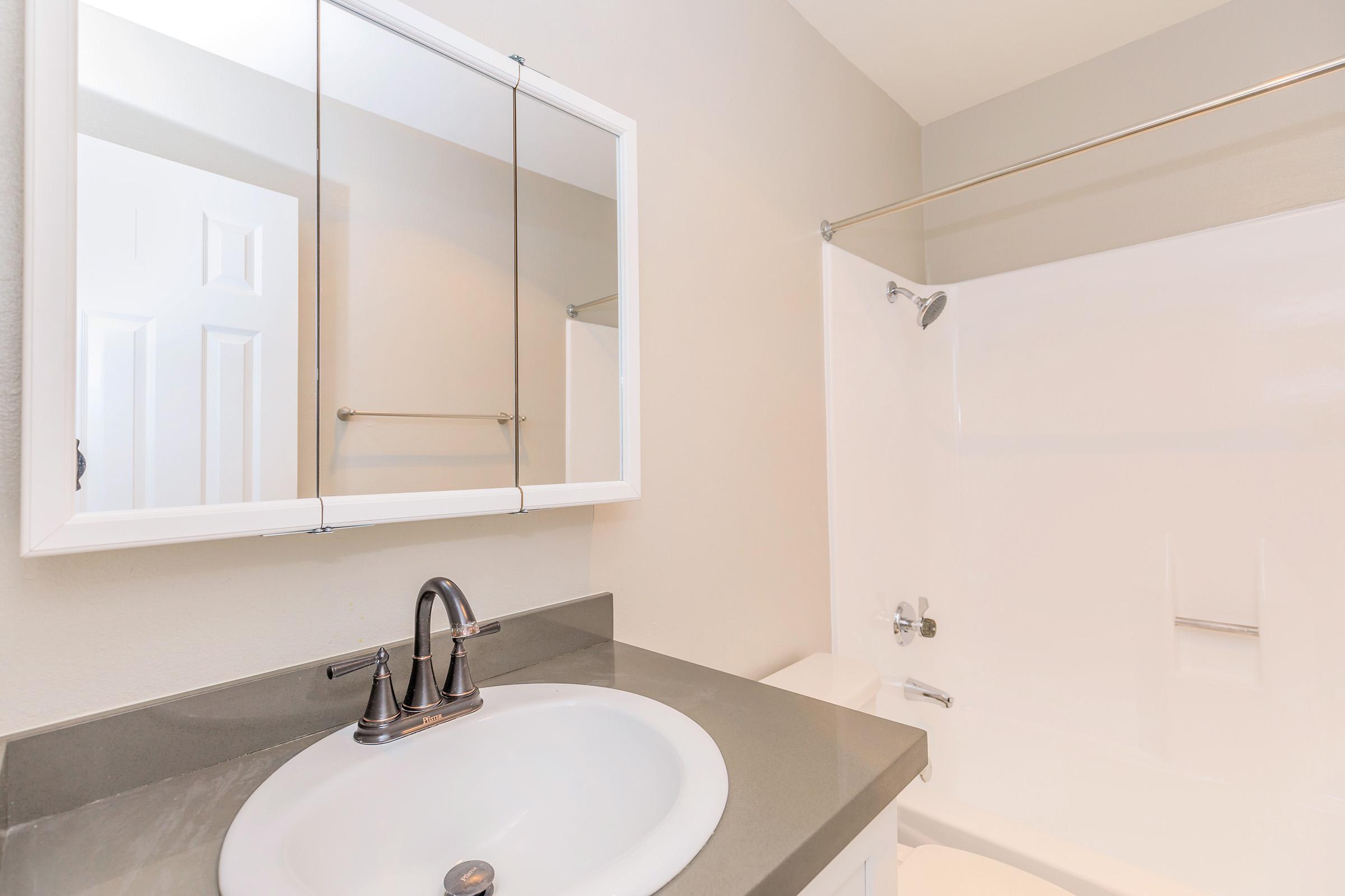 A clean, modern bathroom featuring a double-mirror above a circular sink with a sleek faucet. The shower area has a white tub and a clear shower curtain rod, while a light-colored wall provides a neutral backdrop. A door is slightly visible on the left side.