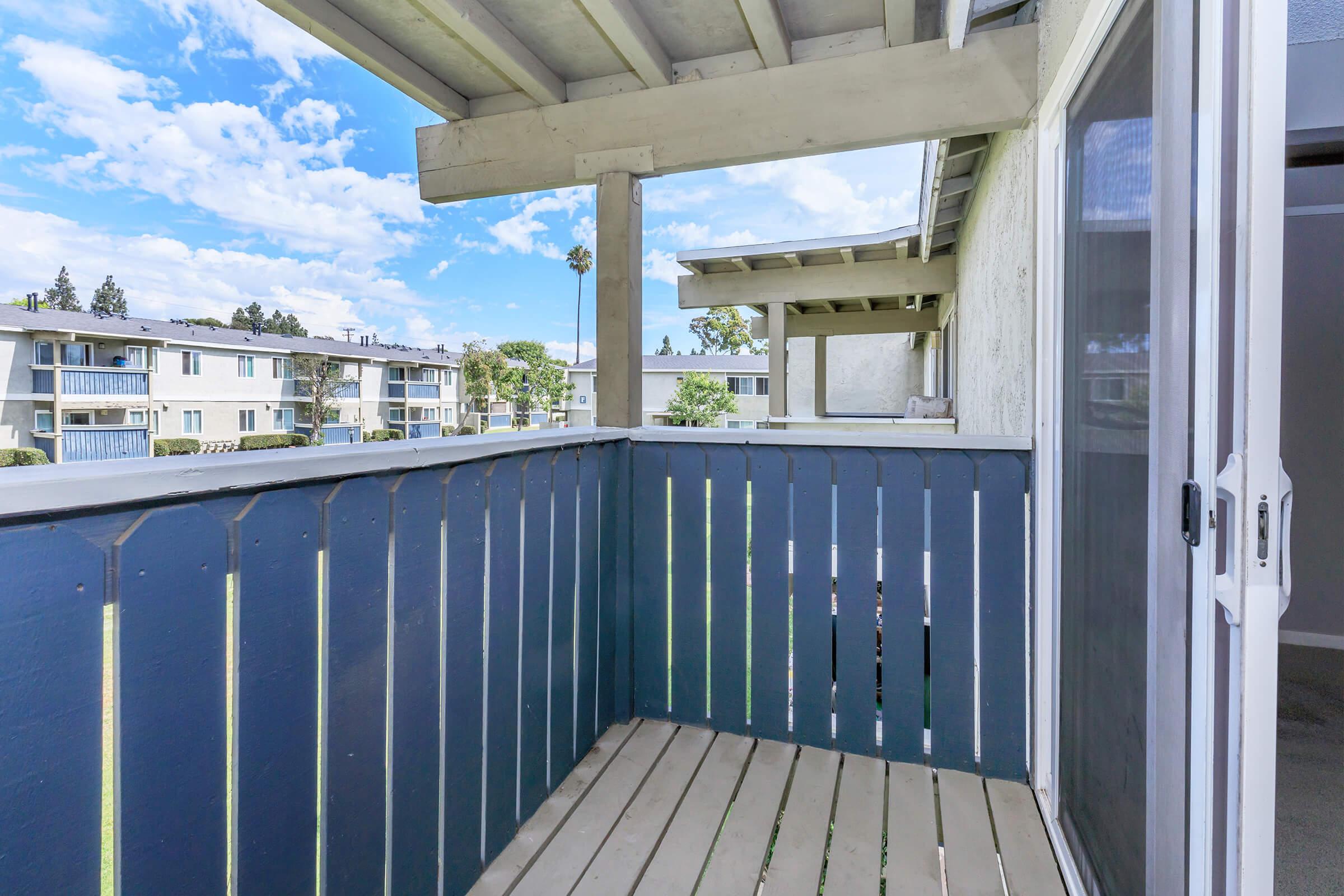 A balcony with wooden flooring and blue-painted railings, overlooking an apartment complex with multiple buildings. The sky is bright with scattered clouds, and there are trees and shrubs visible in the background. The view captures a sense of openness and tranquility.
