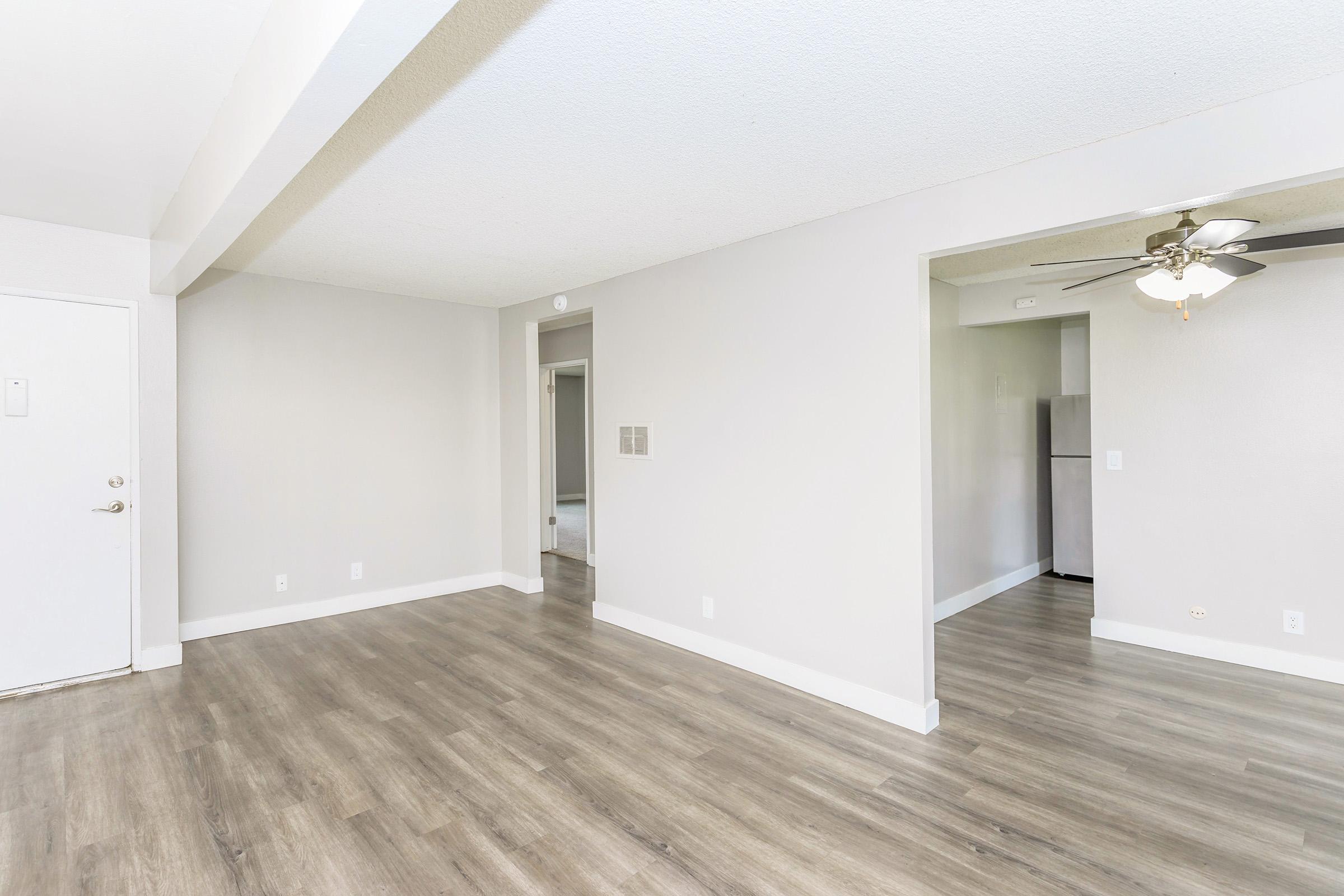 A spacious, light-filled living area featuring light-colored walls and wood-like flooring. A doorway leads to another room, and a ceiling fan is visible in the corner. The overall ambiance is modern and minimalistic, with ample natural light.