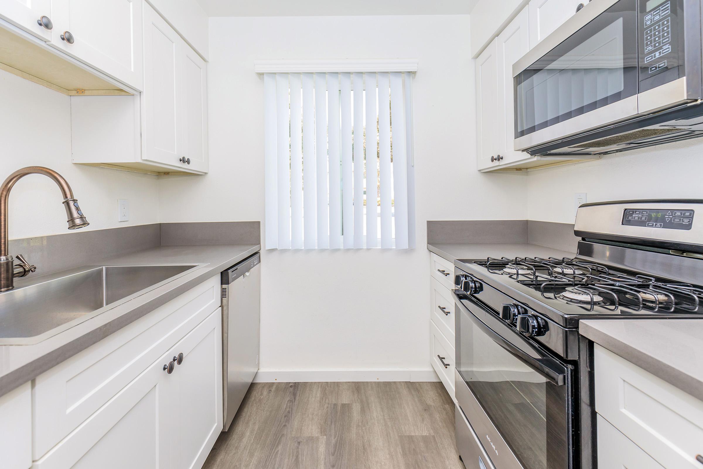 A modern kitchen featuring white cabinetry, a stainless steel sink, a dishwasher, a gas stove, and a microwave. The counter has a gray finish, and there are vertical blinds on the window, allowing natural light to enter the space. The flooring is light-colored laminate.
