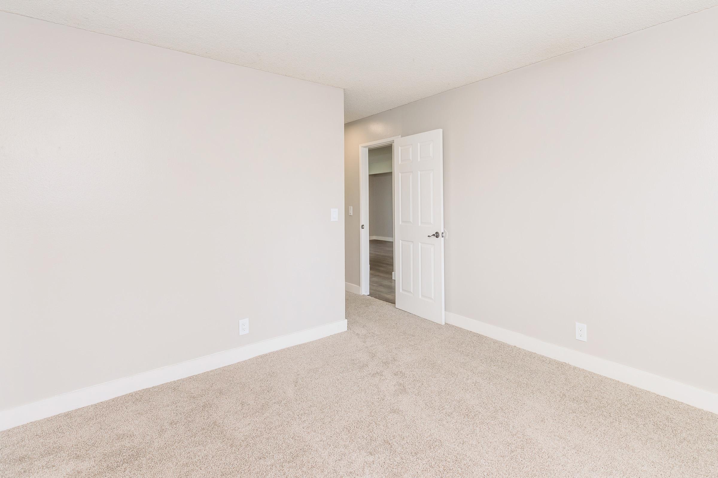 A sparse room with light beige carpet and plain white walls. There's an open door leading to another room, and the space is well-lit, creating a clean and neutral ambiance. No furniture is present, giving it an open and airy feel.