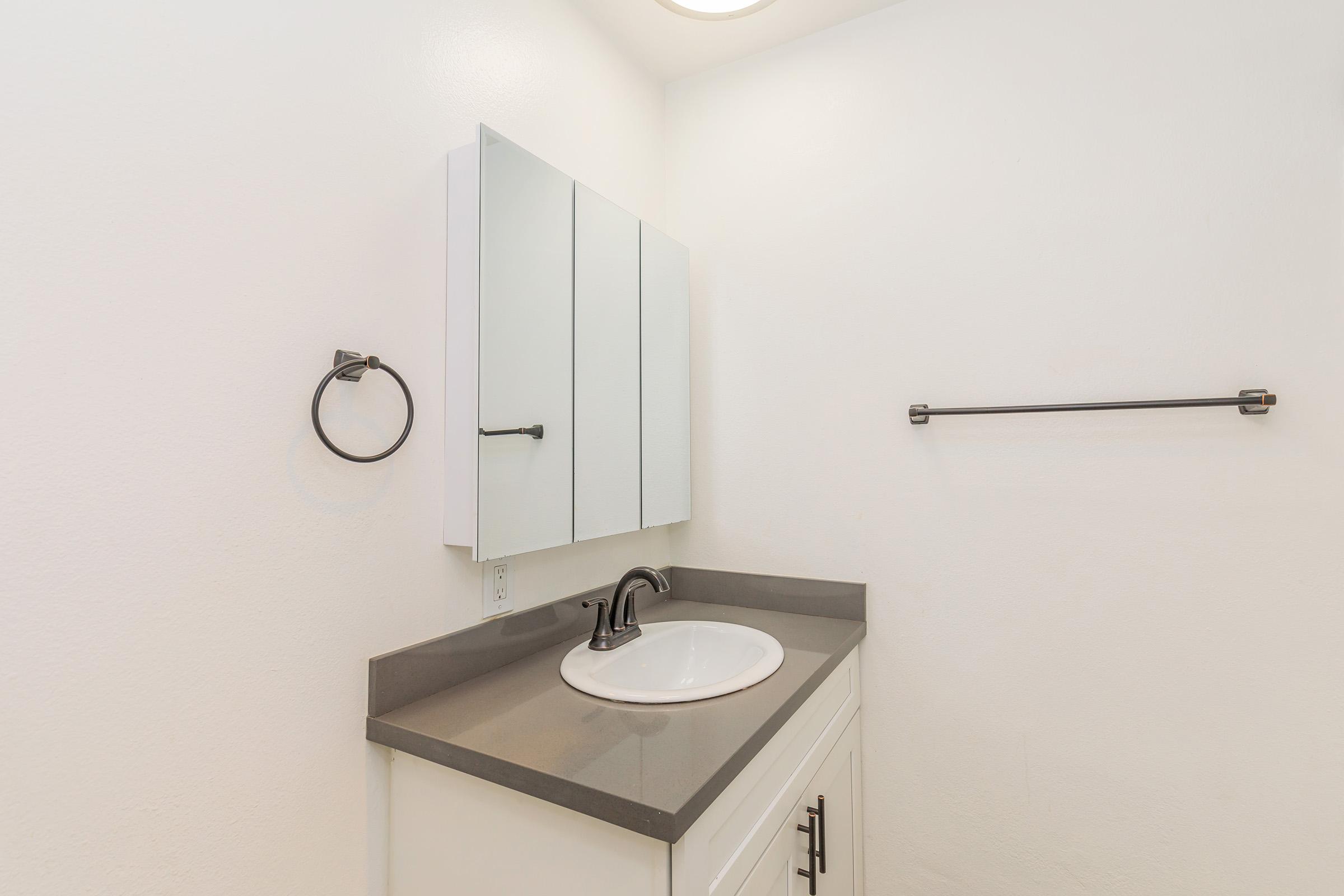A modern bathroom featuring a white sink with a black faucet, a wall-mounted mirror cabinet, and a simple gray countertop. The walls are painted white, and there is a towel ring and a horizontal towel bar on the walls, creating a clean and minimalist appearance.