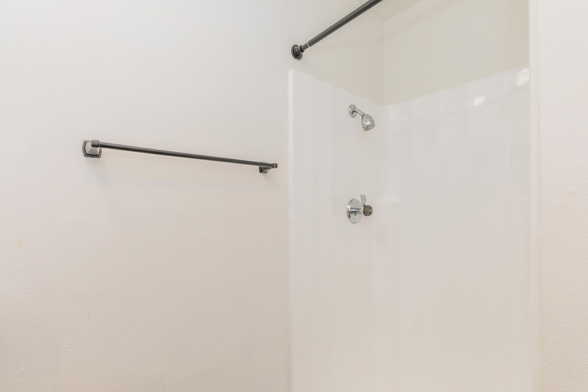 A clean, modern shower area featuring a white shower wall, a chrome showerhead, and two black towel bars mounted on the wall. The overall design is minimalistic with neutral colors, focusing on functionality and simplicity.