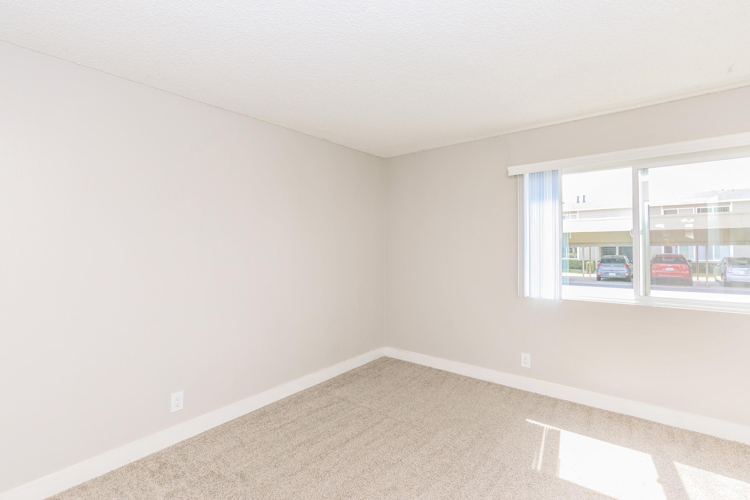 Empty room with light beige walls and carpet. A large window with white blinds lets in natural light, showing a view of parked cars outside. The space appears clean and well-maintained, suitable for moving in or decorating.