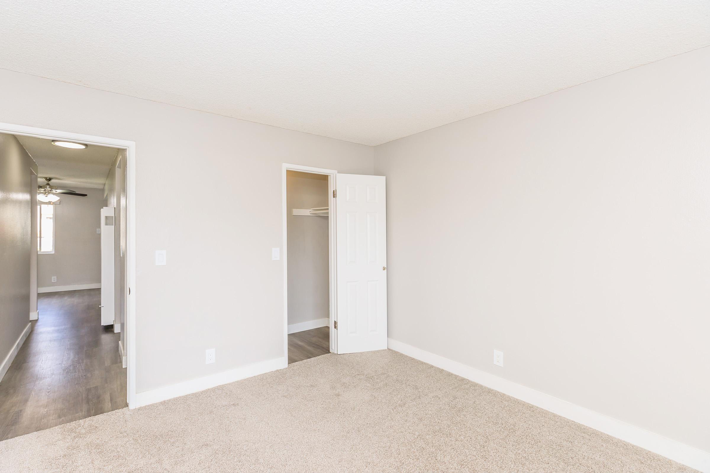 Empty room with light gray walls and carpeted floor. A closed white door on the right leads to a closet, while another door on the left opens to a hallway. The space is well-lit with a ceiling light fixture visible in the background, suggesting a clean and modern living environment.