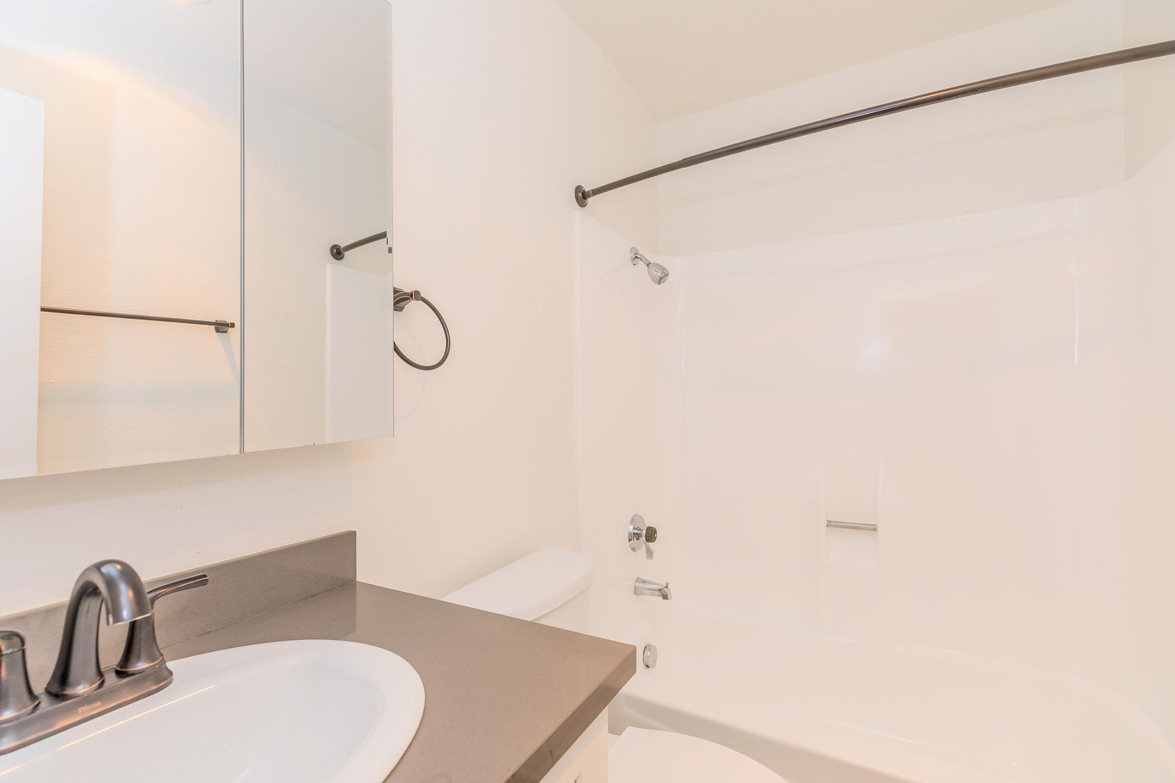 A clean, modern bathroom featuring a white bathtub and shower combination, a sleek black countertop with a sink, and a mirror above the sink. The walls are plain and bright, enhancing the spacious feel, with a black towel bar and minimalistic fixtures. The overall design is simple and contemporary.