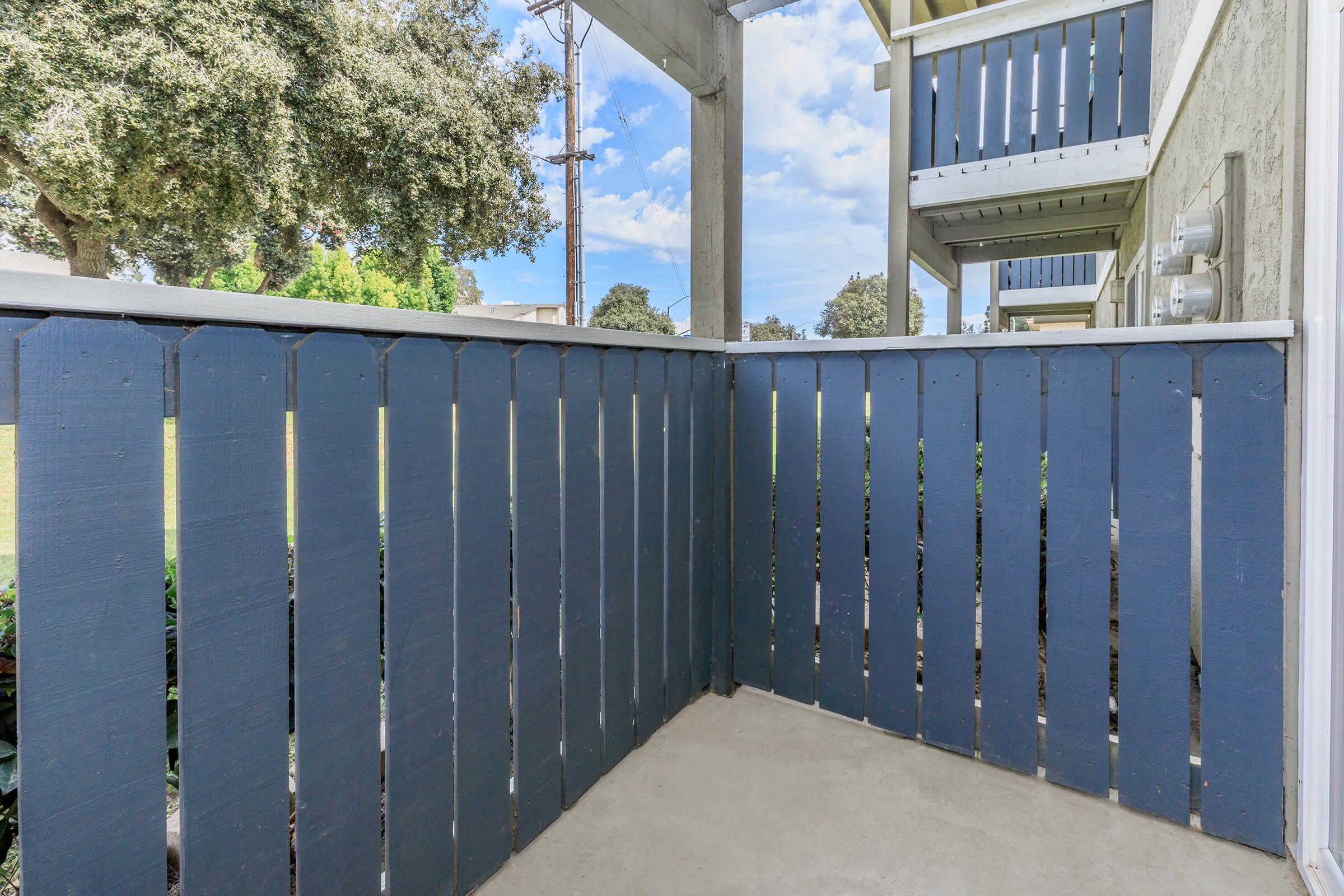 A corner view of a patio enclosed by a blue wooden fence, with a concrete floor. In the background, greenery and a clear blue sky with scattered clouds can be seen, along with a glimpse of a building structure featuring a balcony.