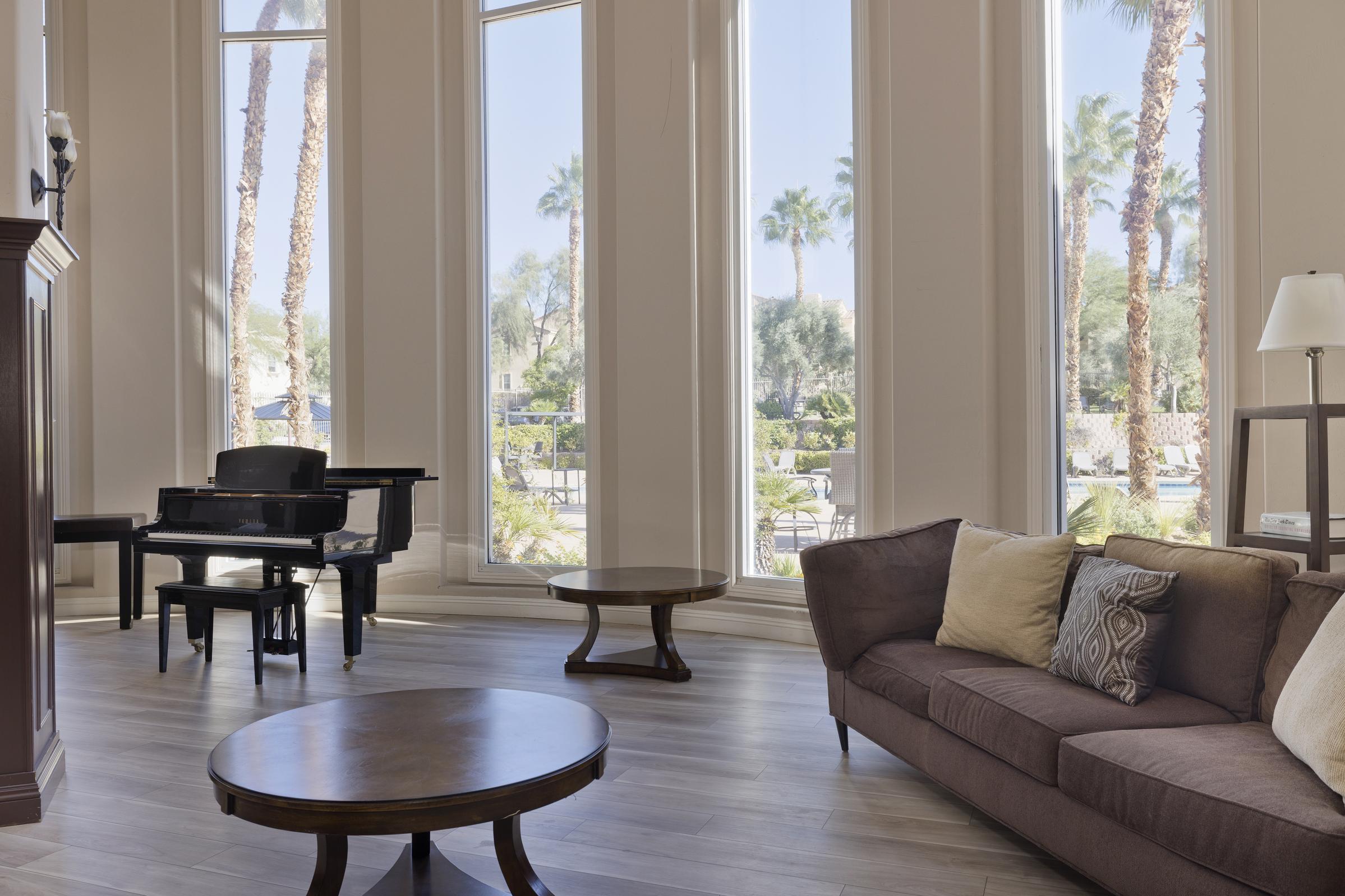 A bright and spacious living area features large windows with views of palm trees outside. A black piano is positioned beside a round wooden coffee table, while a comfortable brown sofa with decorative pillows fills the space. The room has a modern, elegant feel with natural light flooding in.