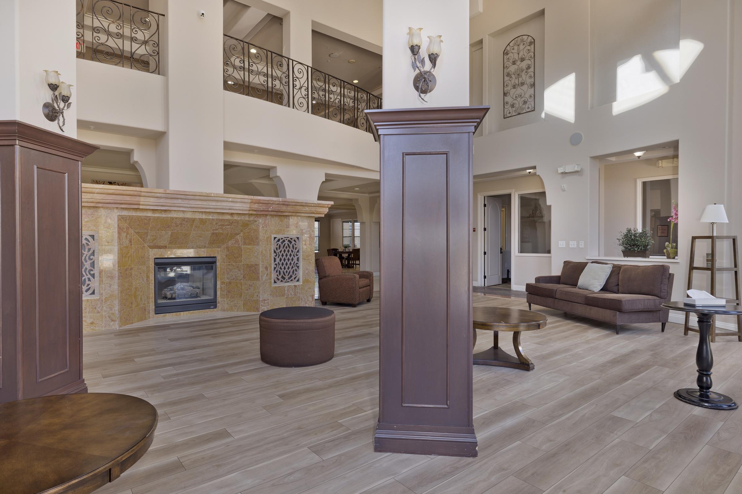 A spacious, well-lit lobby featuring a decorative fireplace, elegant wooden pillars, and modern furnishings. Large windows allow natural light to fill the room, enhancing the inviting atmosphere. Comfortable seating areas include a brown sofa and a round table, creating a cozy and welcoming environment.