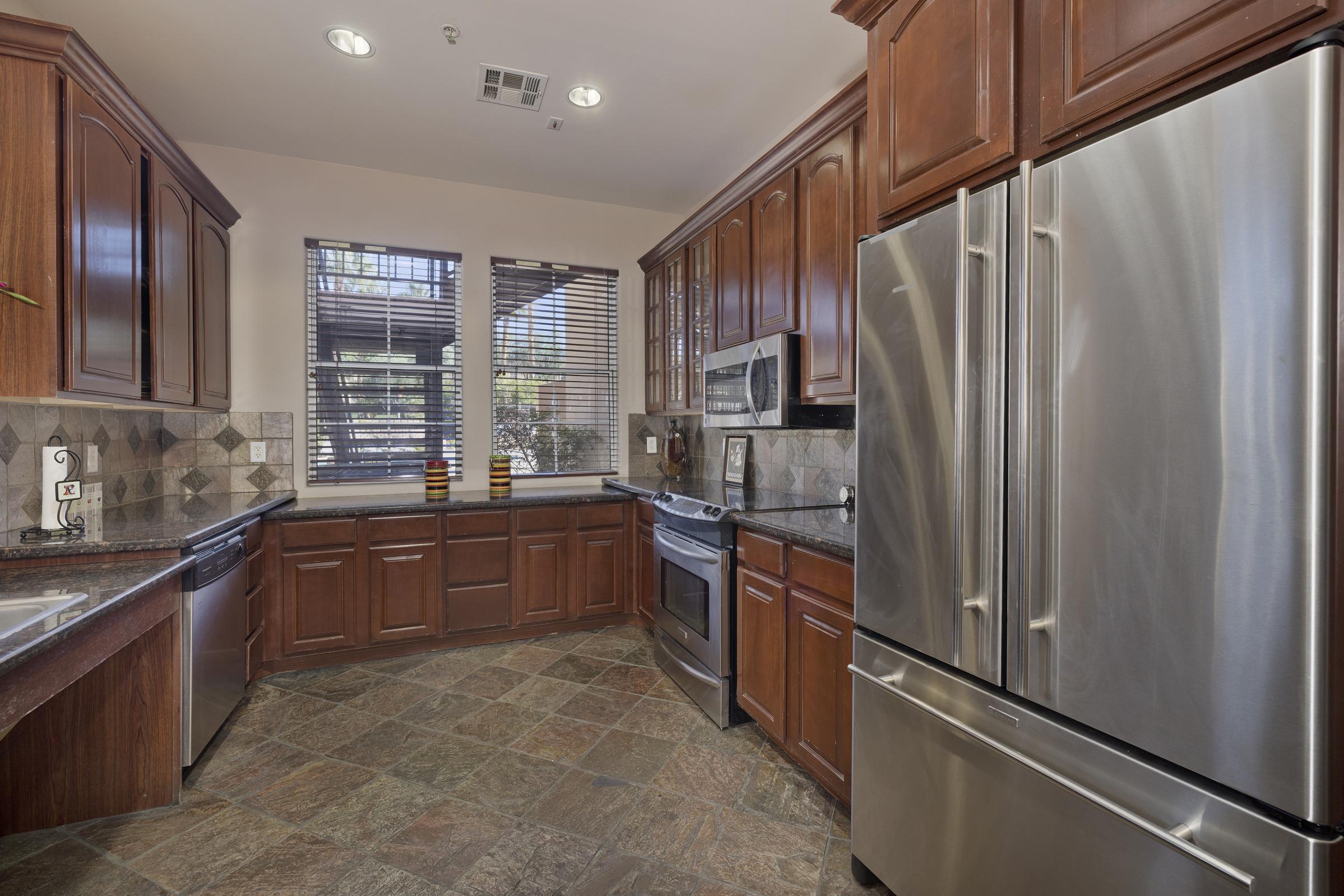 A spacious kitchen featuring dark wooden cabinetry, sleek stainless steel appliances including a refrigerator and microwave, and a textured stone tile floor. Natural light filters through windows with wooden blinds, illuminating the counters and creating a warm, inviting atmosphere.