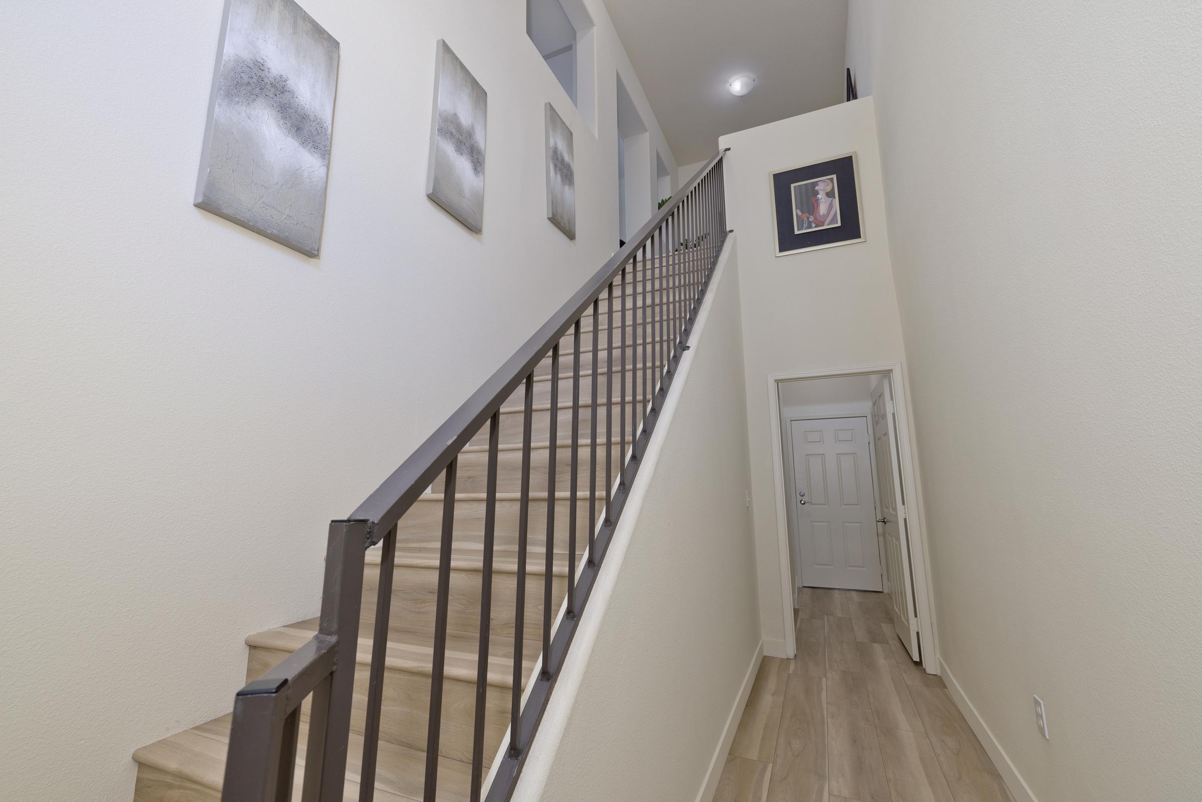 A view of a well-lit hallway featuring a staircase with a metal railing leading upward. The walls are adorned with abstract art pieces, and a door is visible at the bottom, suggesting access to another room. The light wood flooring complements the neutral color scheme of the walls.