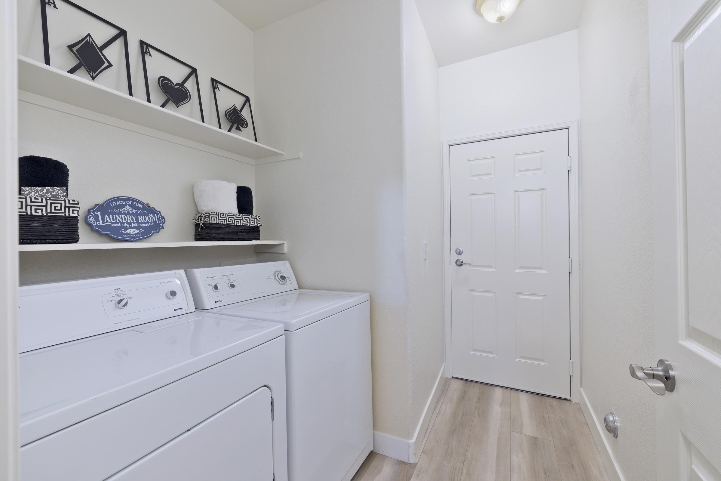 A clean, well-lit laundry room featuring a stacked washer and dryer, a white shelf with neatly folded towels, and decorative wall frames. A door leads to another area, and the flooring is light wood. The overall aesthetic is modern and organized, with a sign indicating "Laundry Room."
