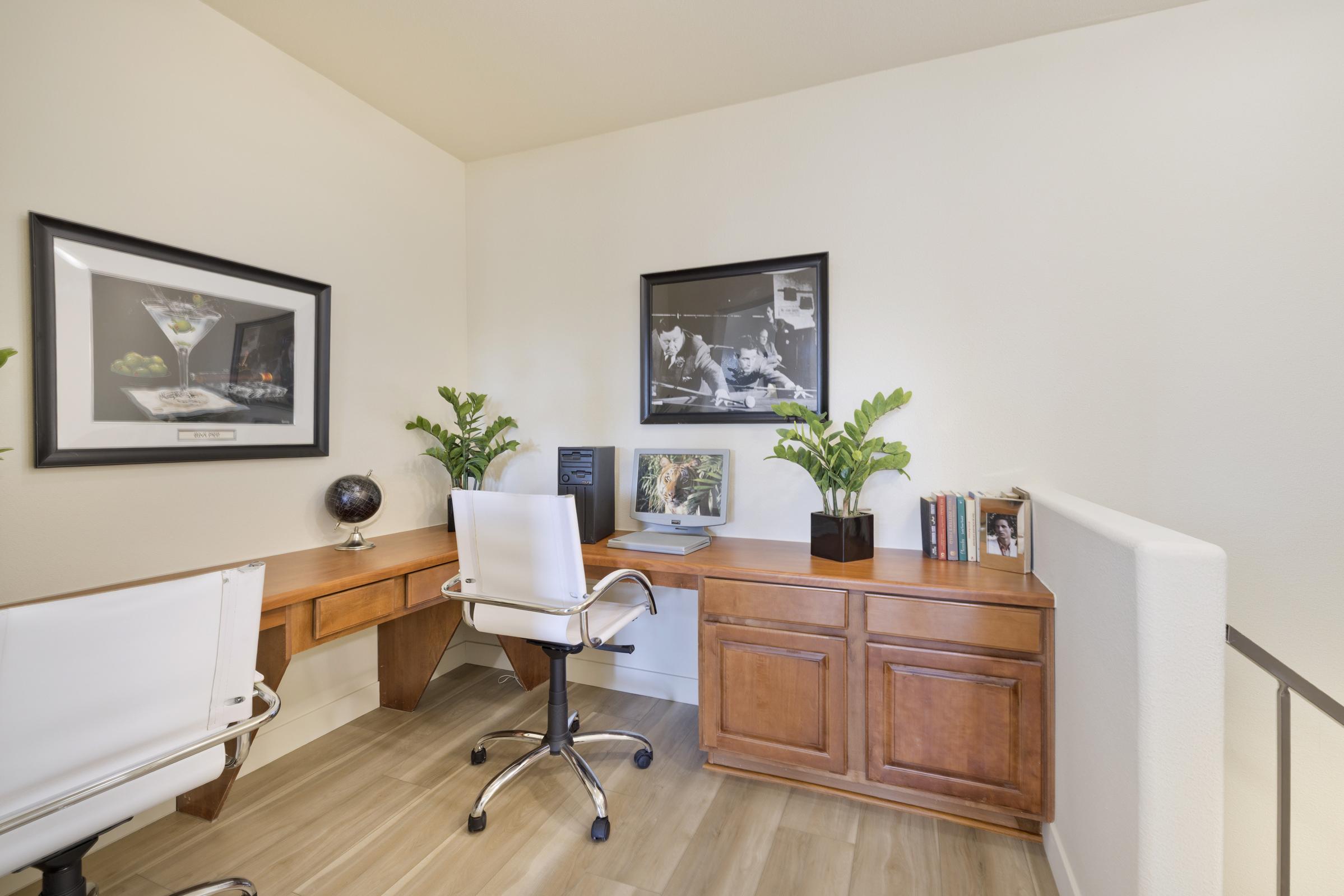 A stylish home office featuring a wooden desk with a computer, two white swivel chairs, and framed black-and-white photographs on the walls. Green plants add a touch of nature, while books are neatly arranged on a shelf nearby. The overall decor is modern and inviting, with a light color scheme.