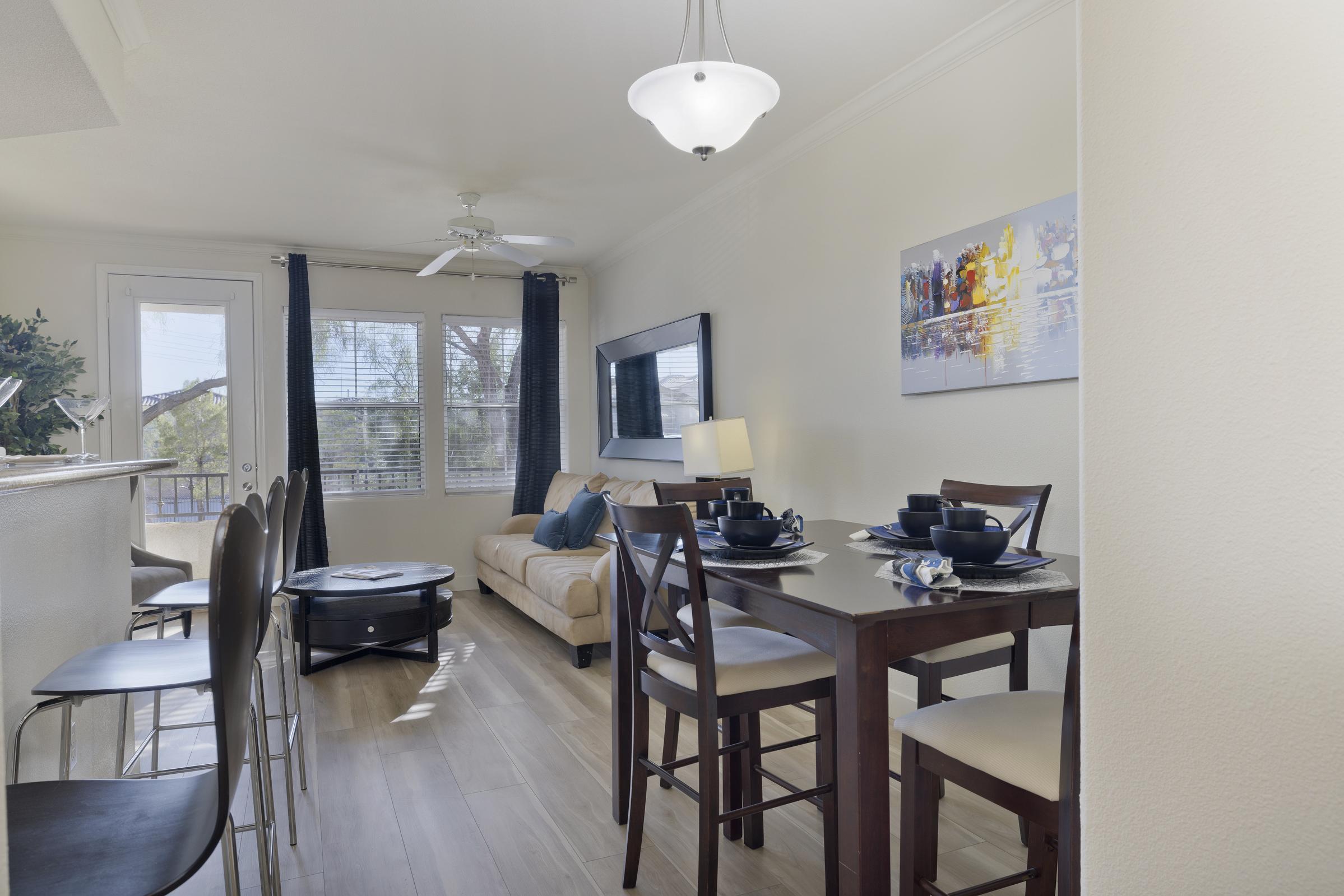 A modern dining area and living space featuring a wooden table set for four, a beige sofa, and a wall-mounted TV. Large windows let in natural light, complemented by light-colored walls. Decorative artwork hangs on the wall, and a ceiling fan adds to the cozy ambiance.