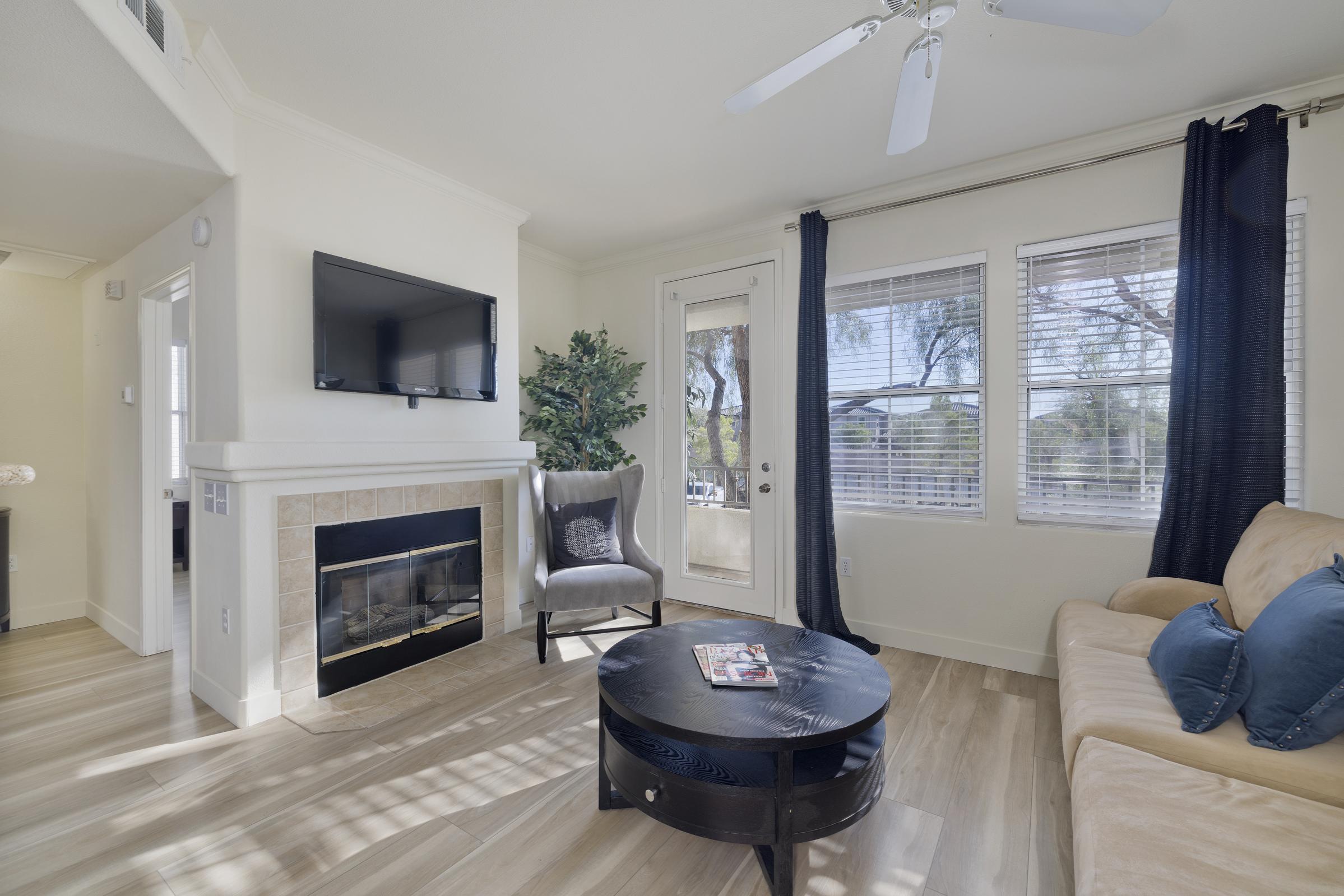 A cozy living room featuring a beige sofa with blue accent pillows, a round coffee table, and a stylish armchair. There's a wall-mounted TV above a modern fireplace, large windows letting in natural light, and a potted plant adding a touch of greenery. The decor is bright and inviting.